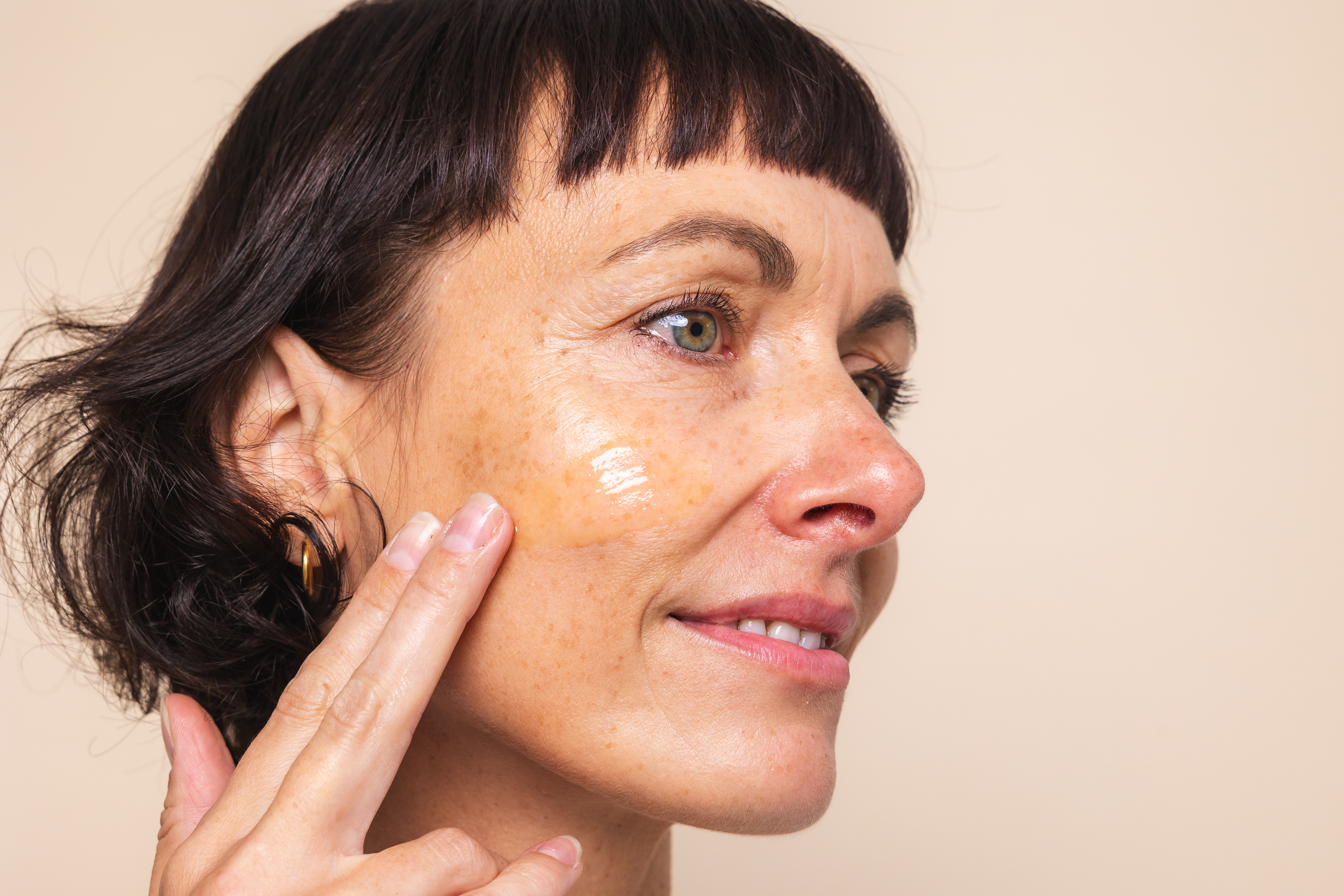 Nahaufnahme einer Frau im Halbprofil, die den Vitamin C Booster von Beyer & Söhne sanft über ihre Wange streicht und mit einem sanften Lächeln und einem entspannten Gesichtsausdruck an der Kamera vorbeischaut