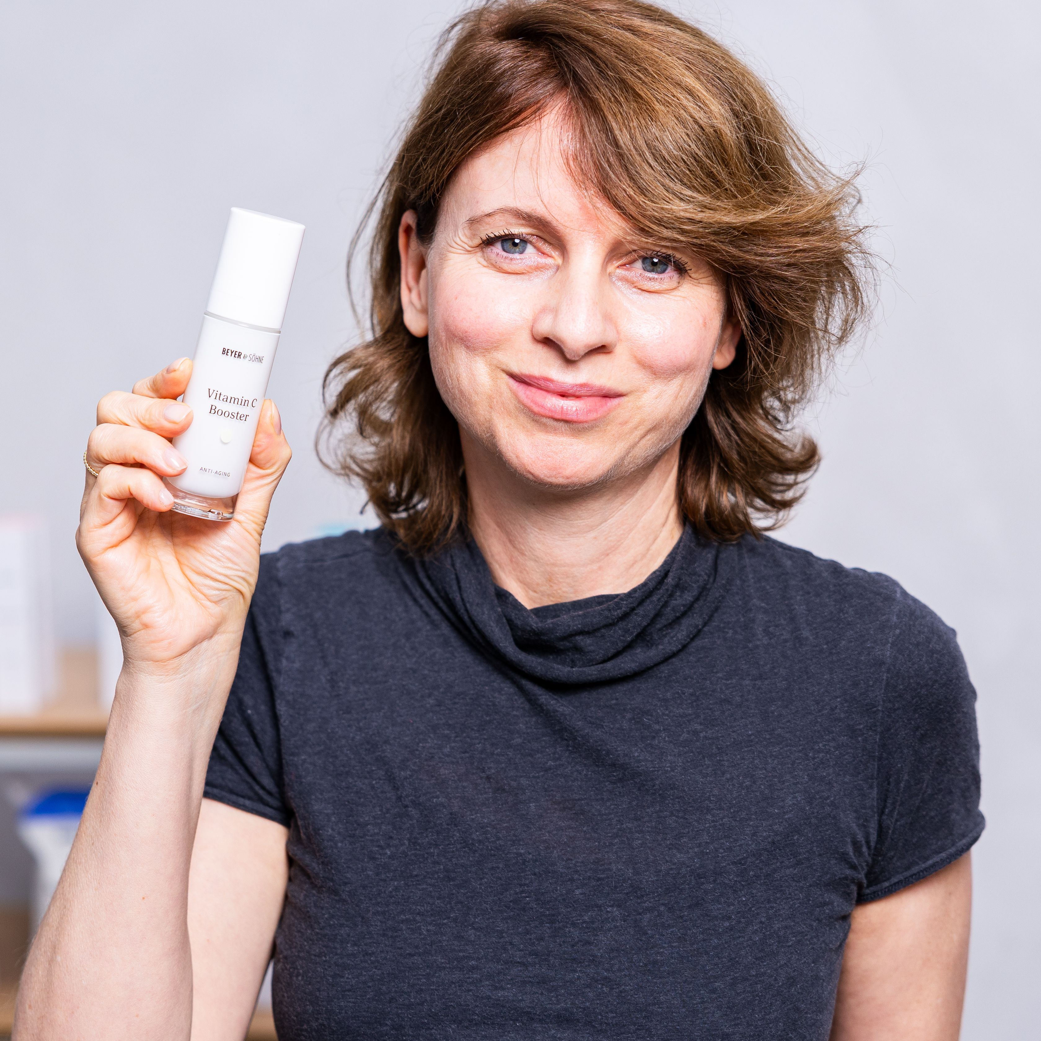 Frau mit kurzen braunen Haaren hält den Vitamin C Booster von Beyer & Söhne in die Kamera. Sie lächelt freundlich und steht vor einem Regal mit weiteren Hautpflegeprodukten der Marke.