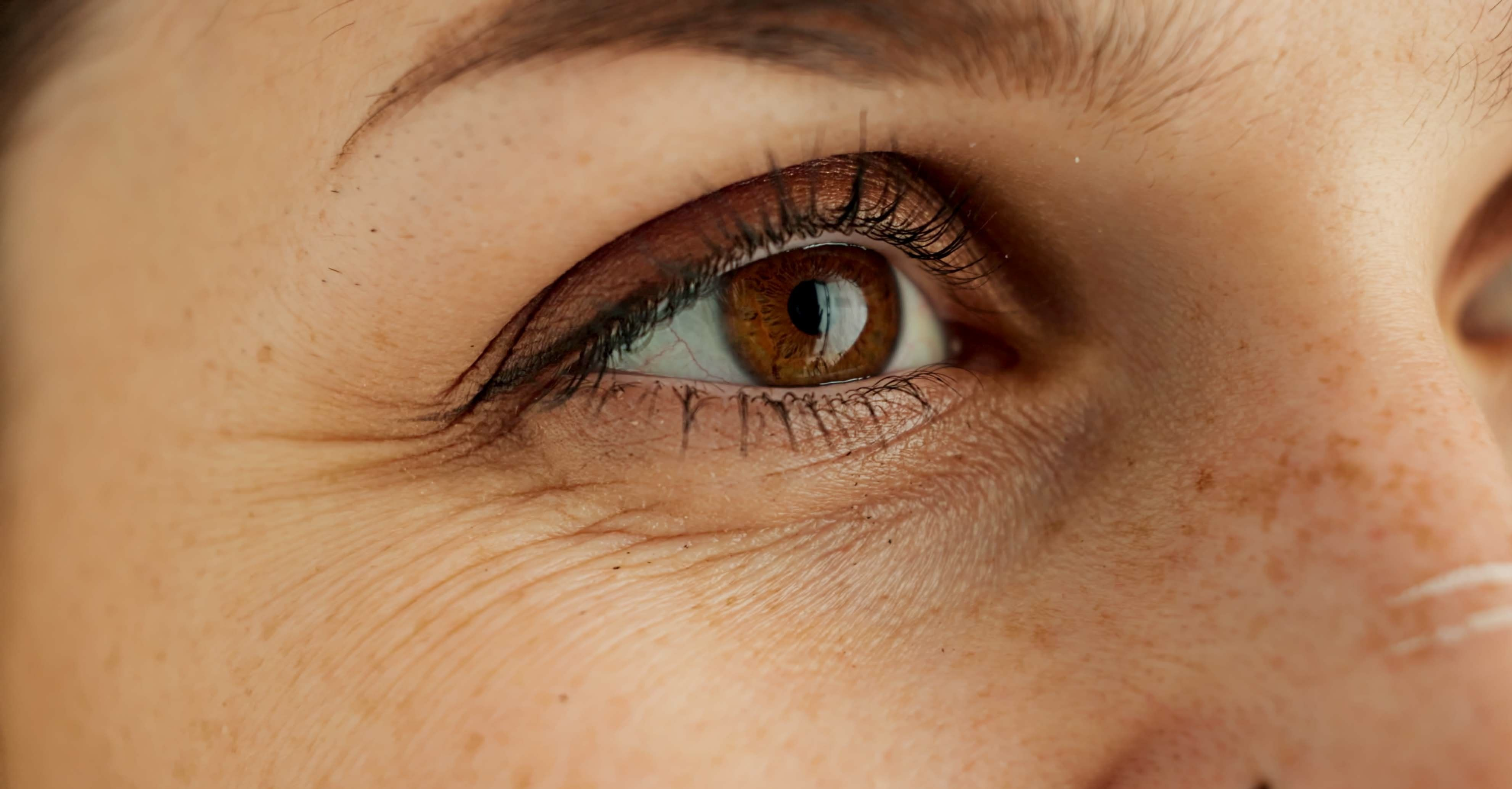Nahaufnahme eines Auges, das an der Kamera vorbeischaut. Kleine Fätchen um das Auge herum zeichnen sich ab und zeigen erste Anzeichen der Hautalterung.