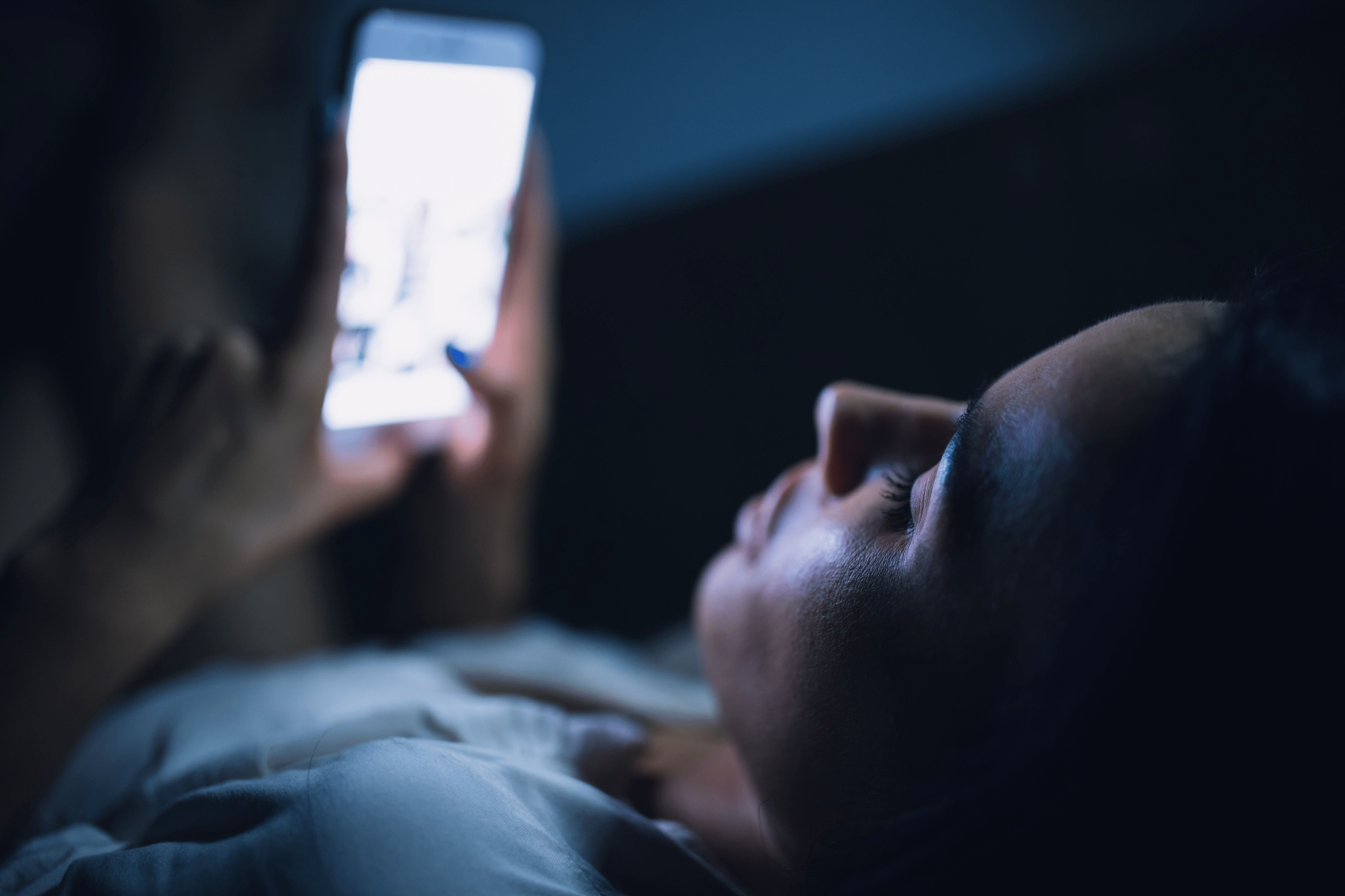 Junge Frau schaut auf ihr Handy in der Dunkelheit. Das blaue Licht des Displays spiegelt sich auf ihrer Gesichtshaut.