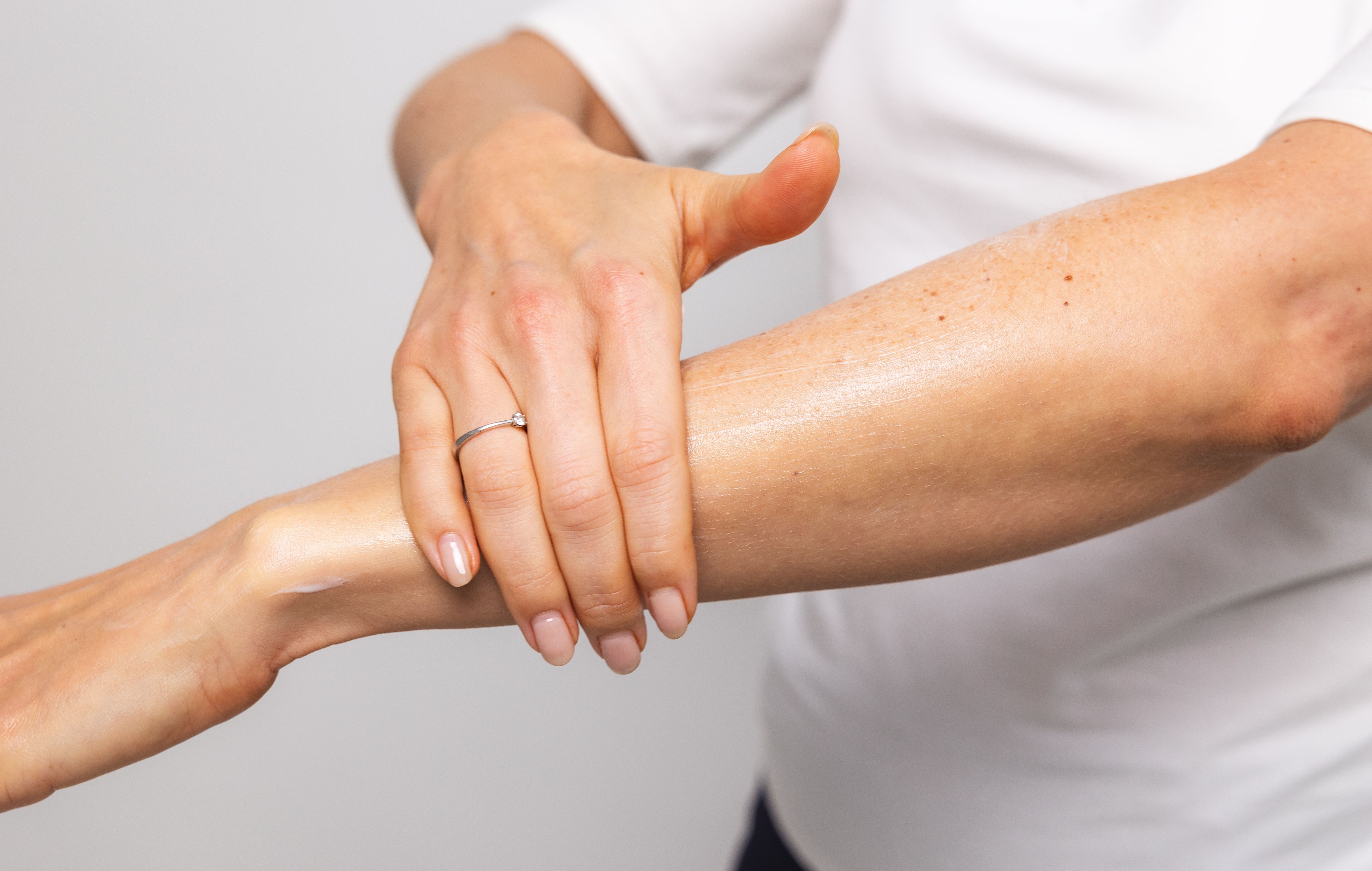 Frau Cremt ihren linken Unterarm sanft mit der rechten Hand ein. Das Foto vermittelt Ästhetik und Sanftheit, durch die zarte Berührung, schöne, natürliche Fingernägel der Frau und einen dezenten Ring. 