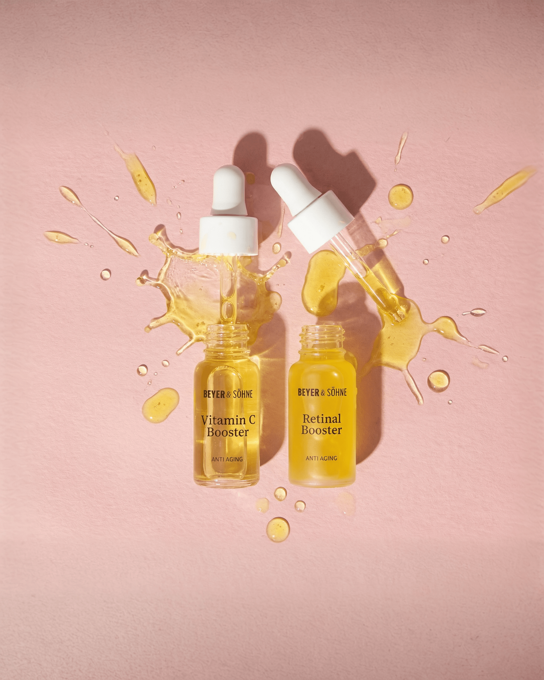 Beyer und Soehne Vitamin C und Retinal Booster. Hochwirksame Anti-Aging Seren mit Pipette auf rosa Untergrund, goldene Textur sichtbar verteilt.