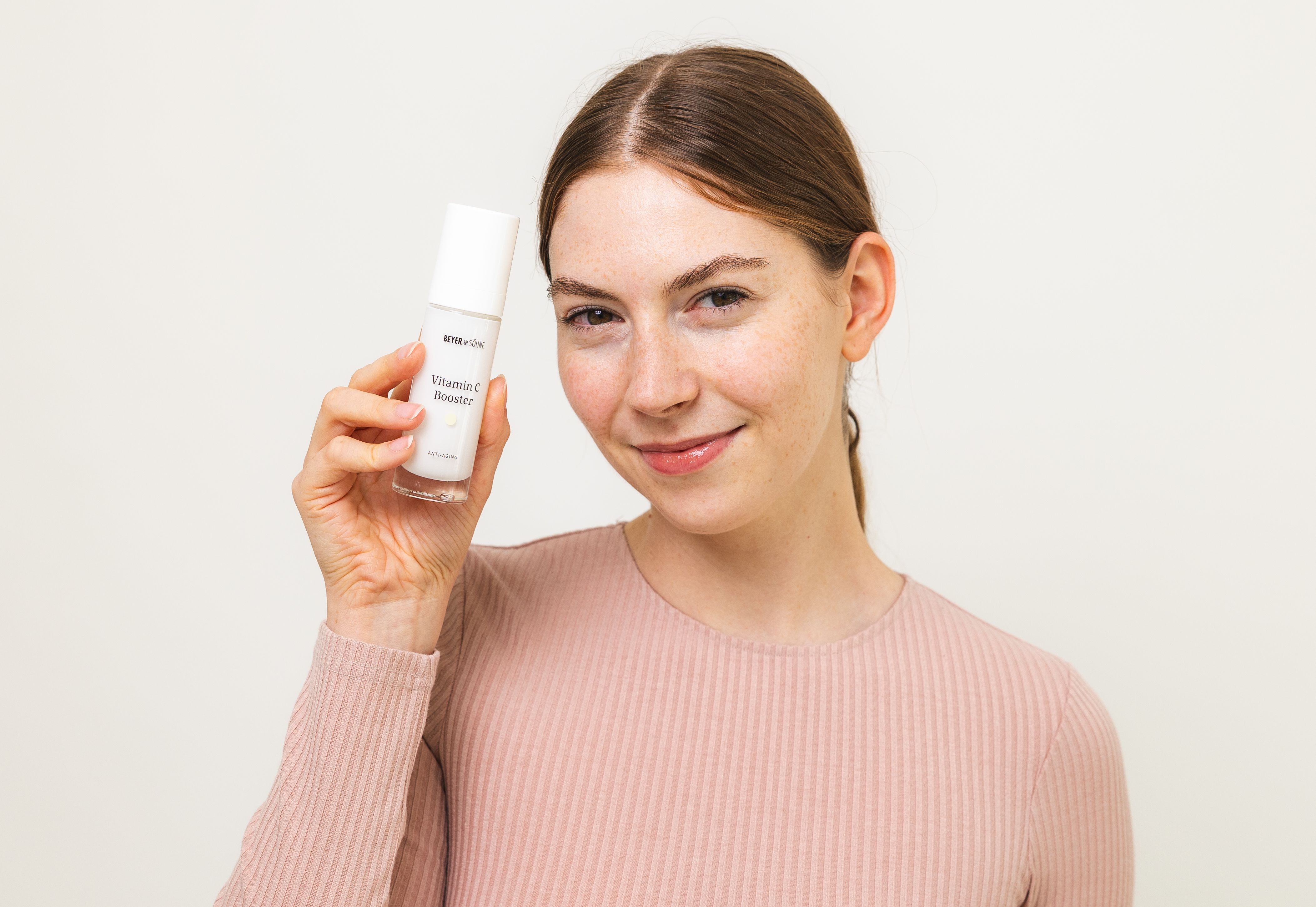 Junge natürliche und strahlende Frau hält den Vitamin C Booster neben ihr Gesicht und schaut mit einem breiten Lächeln in die Kamera