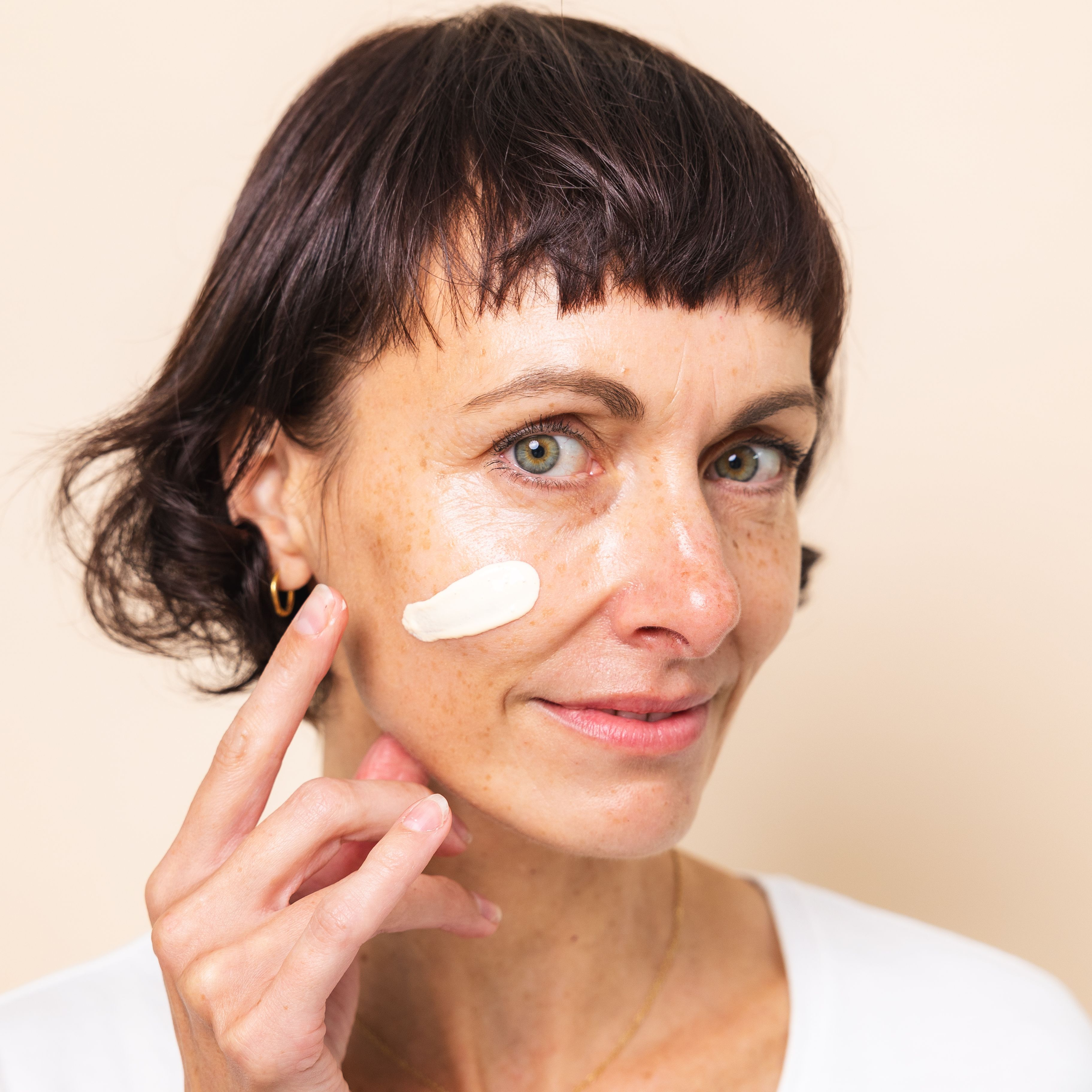 Natürliche Frau Anfang 40 mit kurzem, dunkelbraunen Haar blickt in die Kamera und trägt sich die Hautcreme+ von Beyer & Söhne aufs Gesicht auf