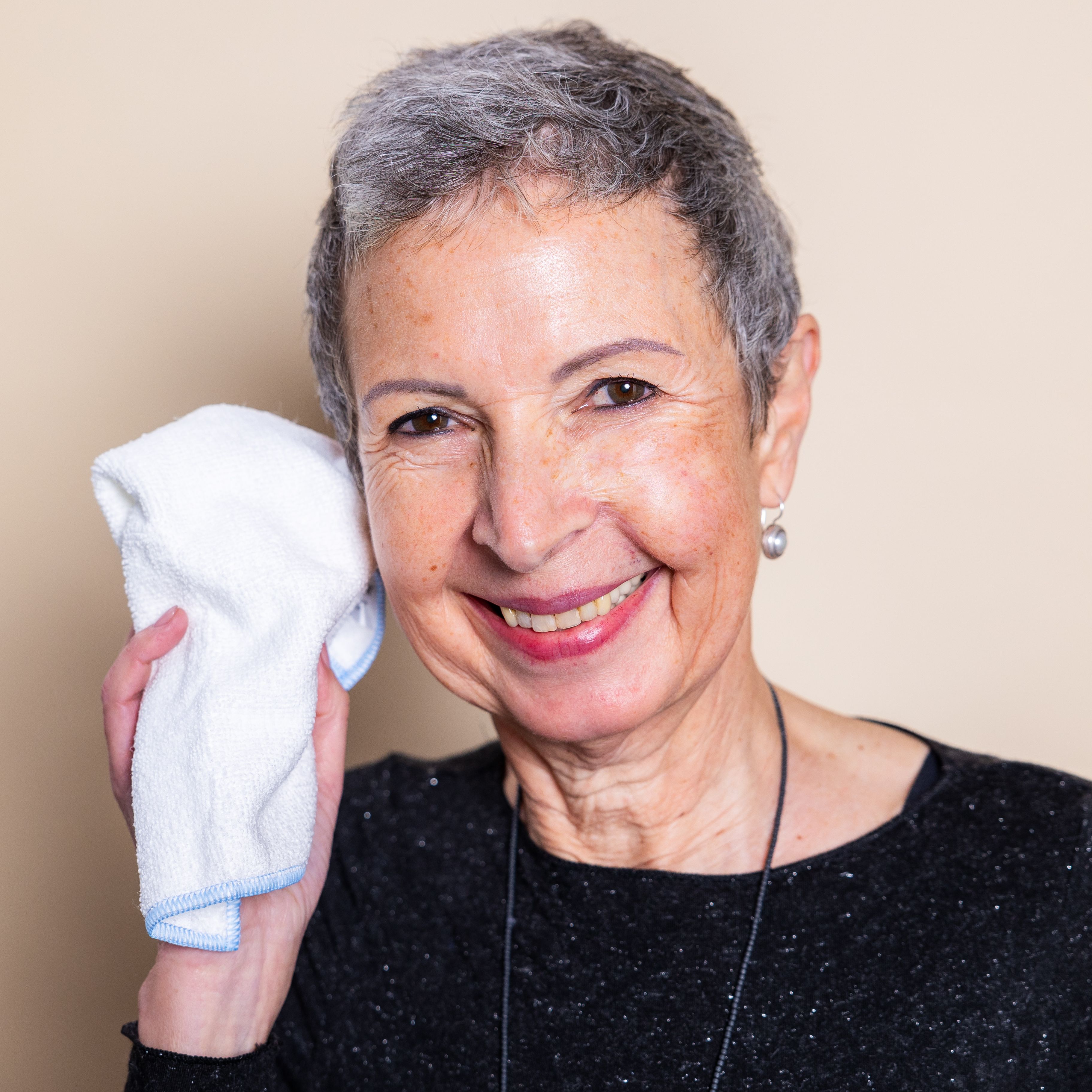 Natürliche Frau Ende 50 mit stylischem Kurzhaarschnitt lächelt strahlend in die Kamera und hält das Mikrofasertuch von Beyer & Söhne an ihr Gesicht