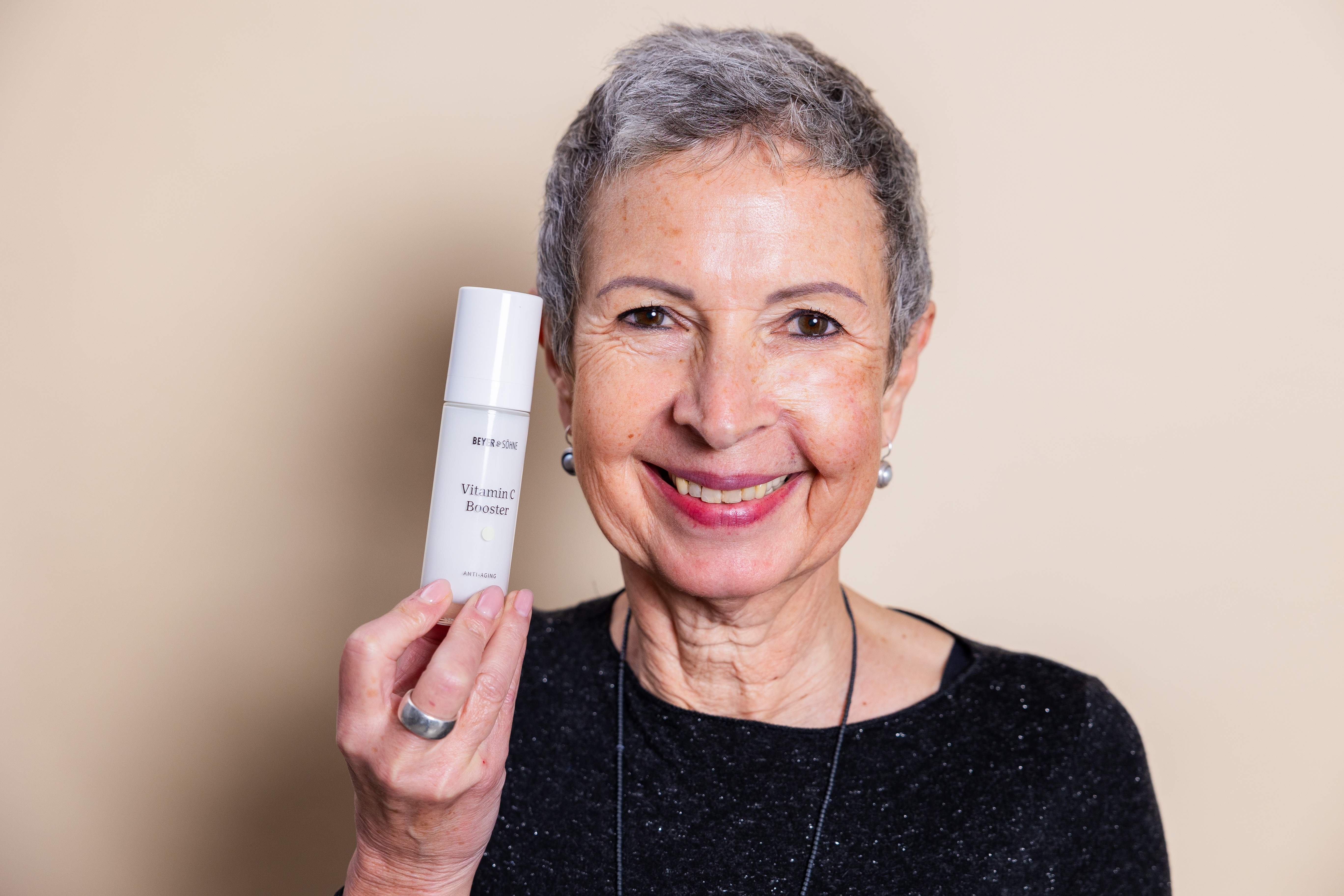 Eine strahlend, natürliche Frau Ende 50, mit einem modernen Kurzhaarschnitt hält den Vitamin C Booster lächelnd in der Hand, während sie frontal in die Kamera schaut.