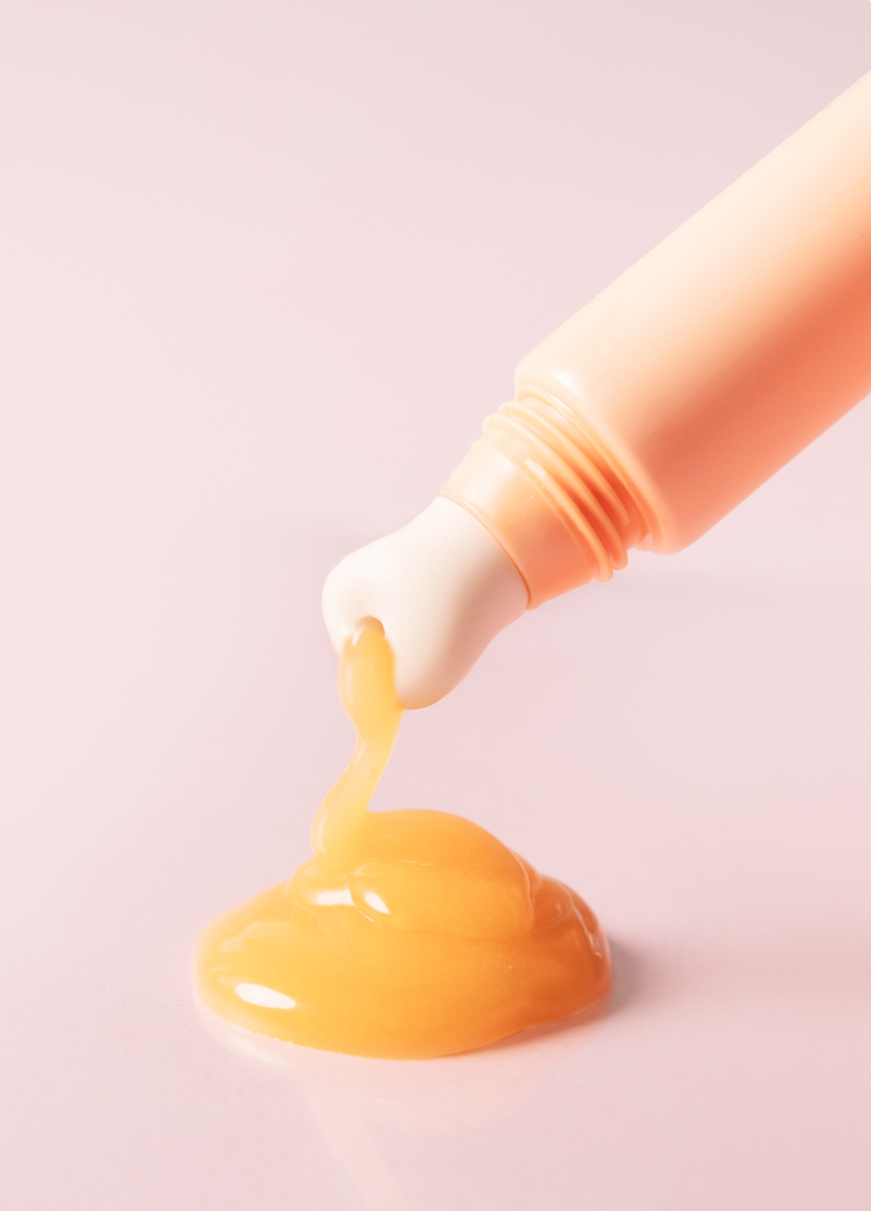 Dayshade Lip Balm SPF 50+ mit Keramik Applikatorspitze, aus der ein Tropfen goldener Balsam fließt, auf zartem rosa Hintergrund.