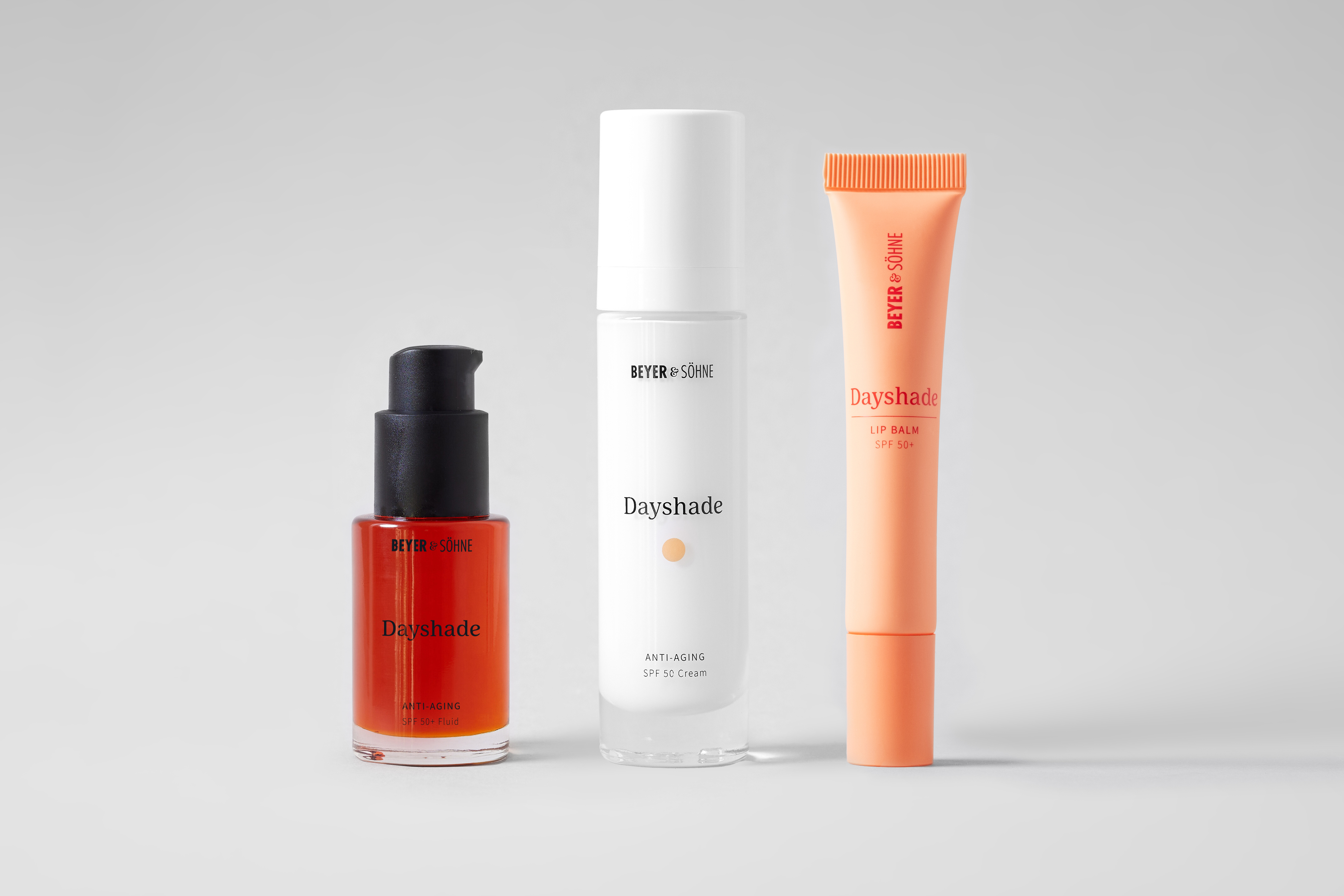 Die drei Beyer & Söhne Dayshade-Produkte nebeneinander: rotes Dayshade Fluid SPF 50+ mit schwarzem Pumpspender, Dayshade Cream SPF 50 in einem weißen Airless-Spender und Lip Balm SPF 50+ in apricotfarbener Tube.