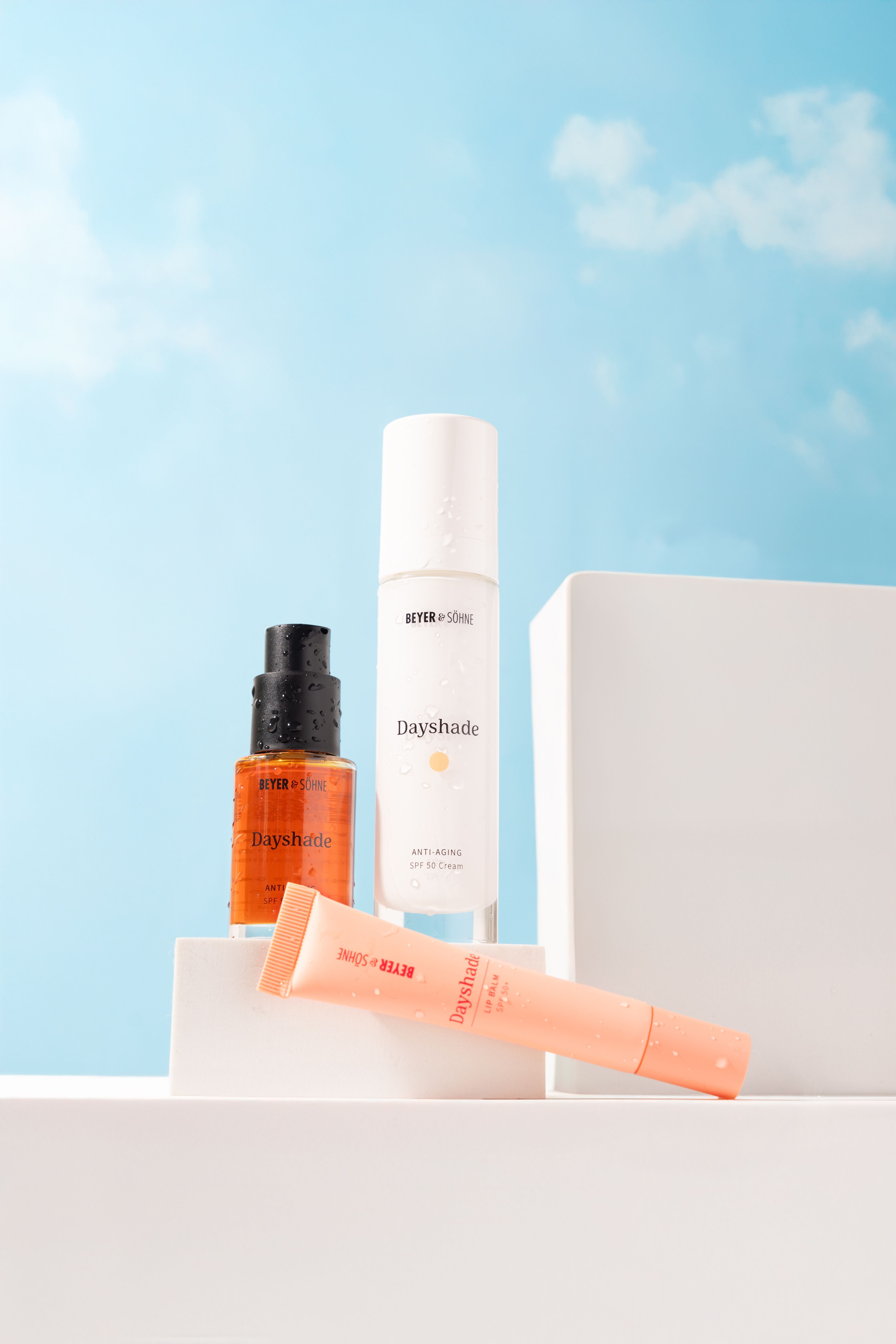 Die drei Beyer & Söhne Dayshade-Produkte vor blauem Himmel: rotes Dayshade Fluid SPF 50+ mit schwarzem Pumpspender, Dayshade Cream SPF 50 in einem weißen Airless-Spender und Lip Balm SPF 50+ in apricotfarbener Tube.