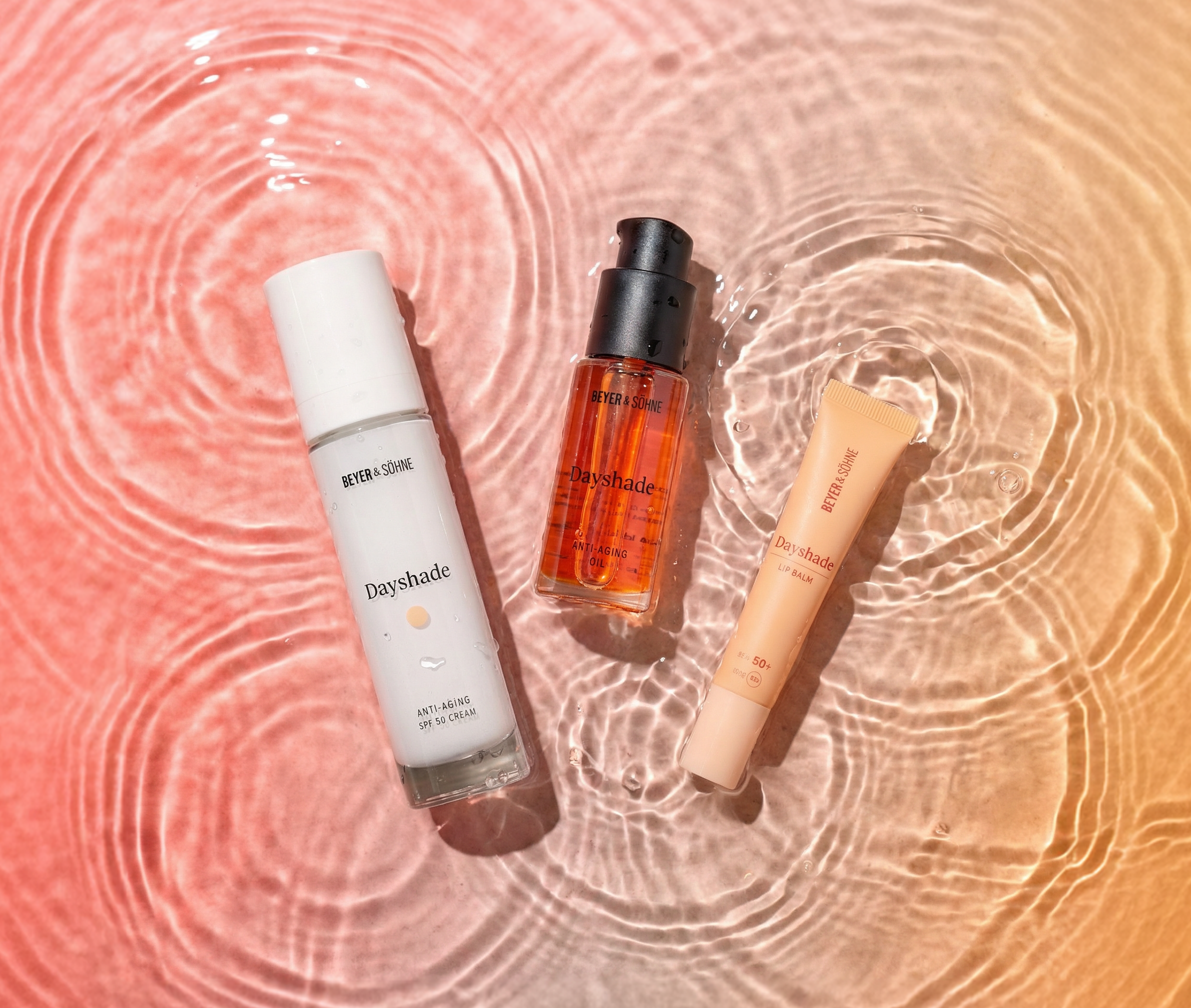 Drei Dayshade Produkte liegen auf einer Wasseroberfläche mit sanften Wellen, umgeben von warmen Farbverläufen in Orange- und Rosétönen, die eine leichte, frühlingshafte Atmosphäre vermitteln.