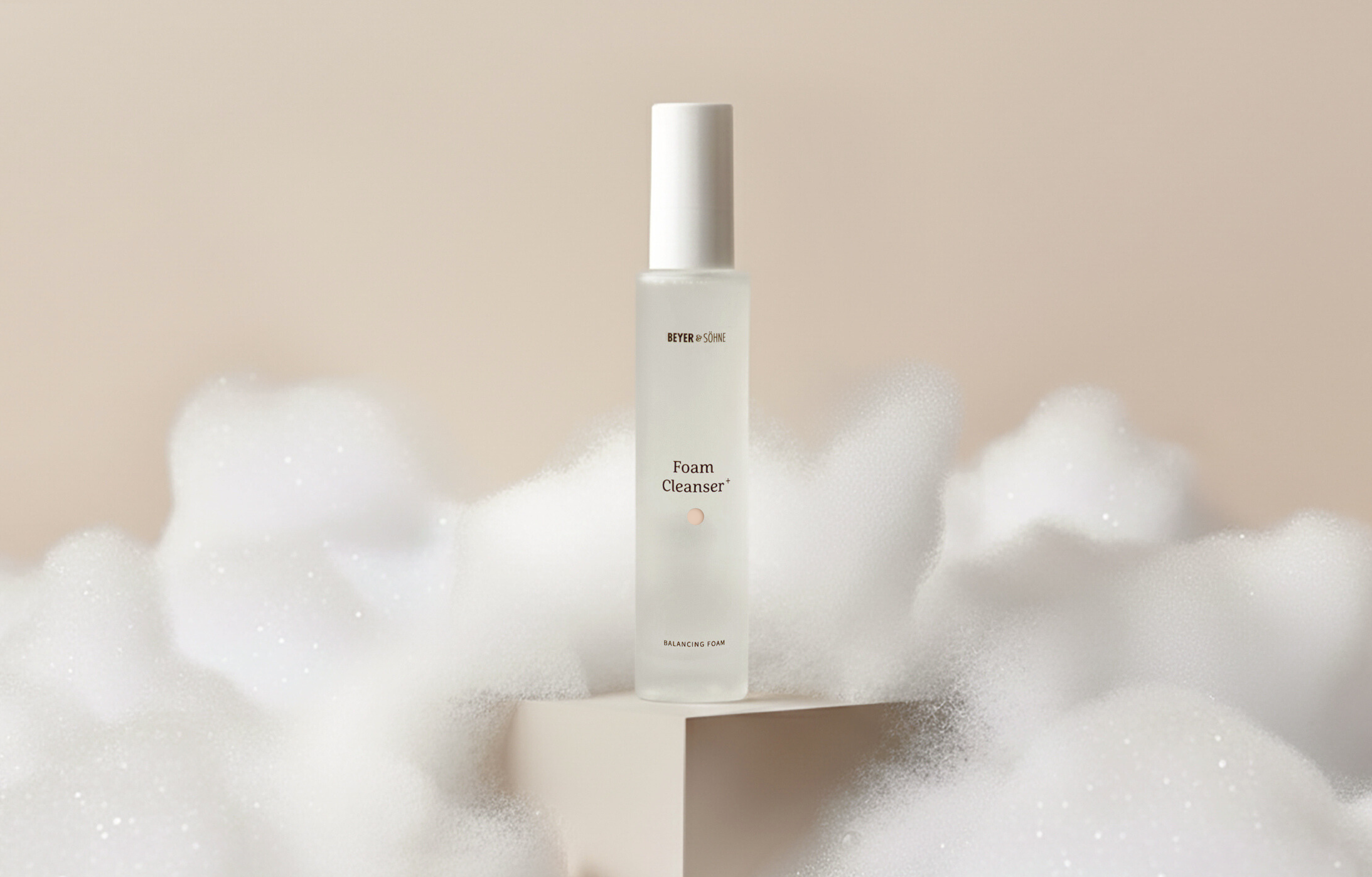 Foam Cleanser steht auf einem kleinen Podest vor warmen, beigen Hintergrund umgeben von feinsten, cremigen Schaum, der wie eine weiche Wolkendecke aussieht.