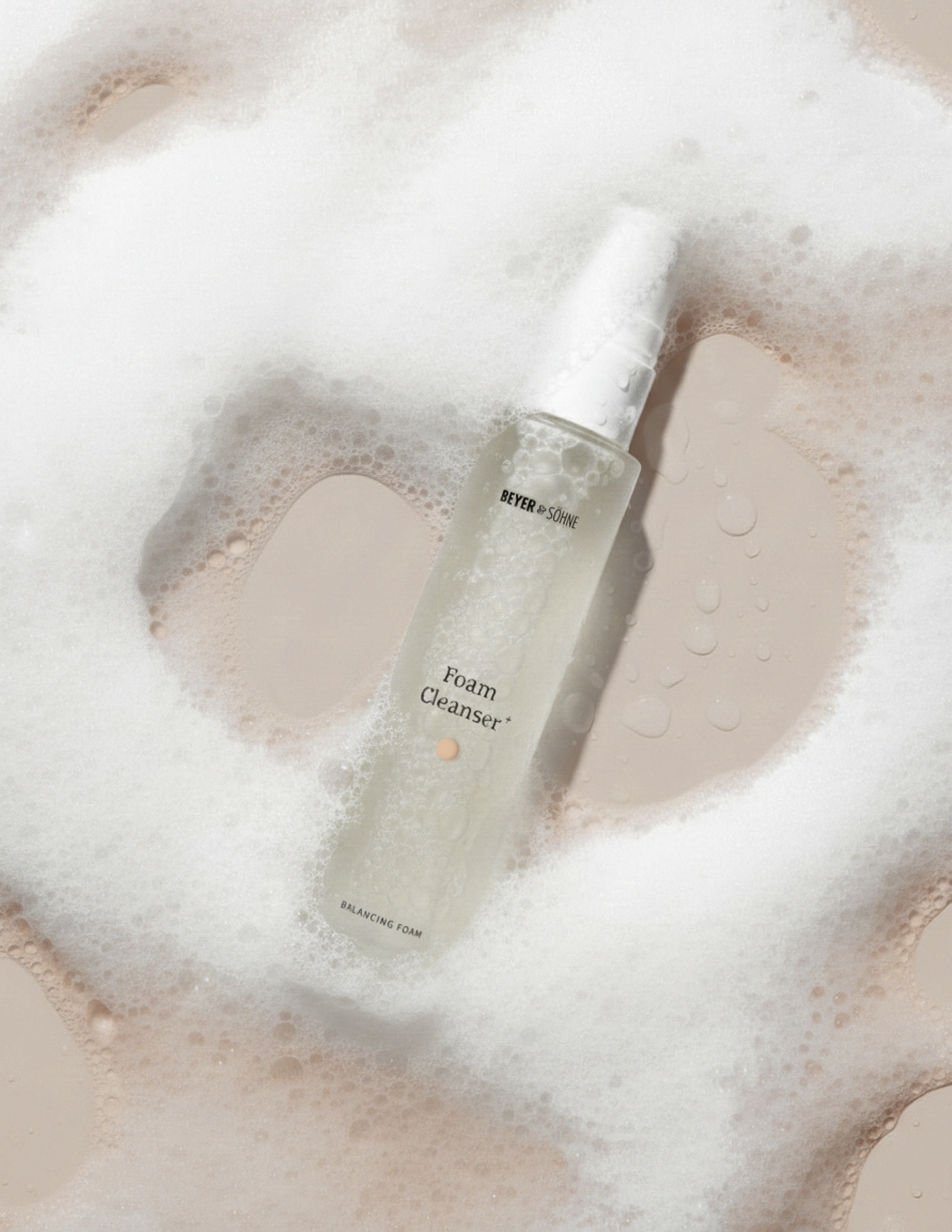 Milchig transparenter Pumpspender mit Foam Cleanser+ von Beyer & Söhne eingebettet in feinen weißen Schaum mit einigen Wassertropfen drumherum. 