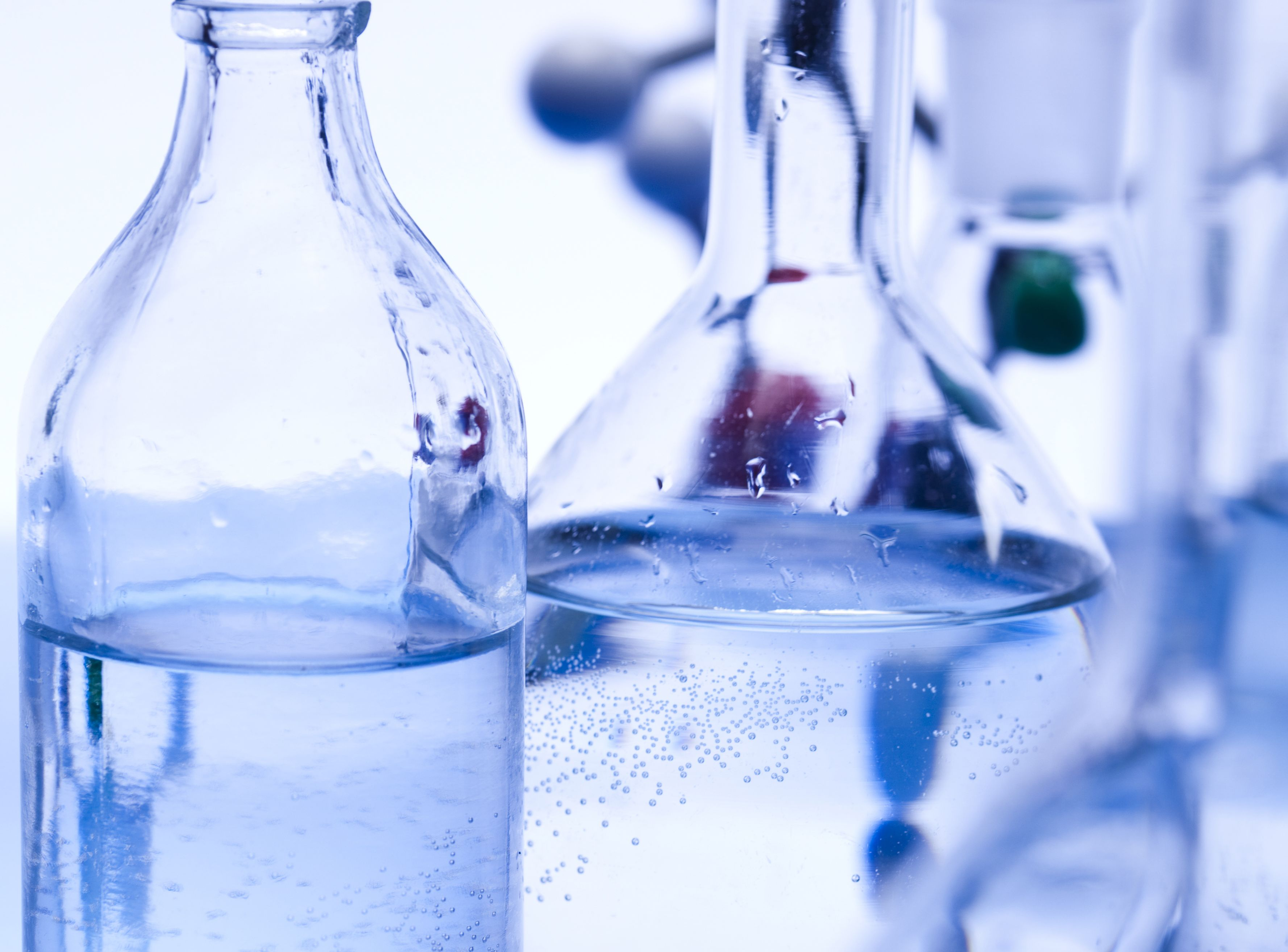 Mehrere Glasbehälter, die man aus dem Labor kennt, stehen mit einer klaren Flüssigkeit gefüllt nebeneinander. Das Glas der Behälter ist leicht kornblumenblau gefärbt und vermittelt eine laborartige Atmosphäre.