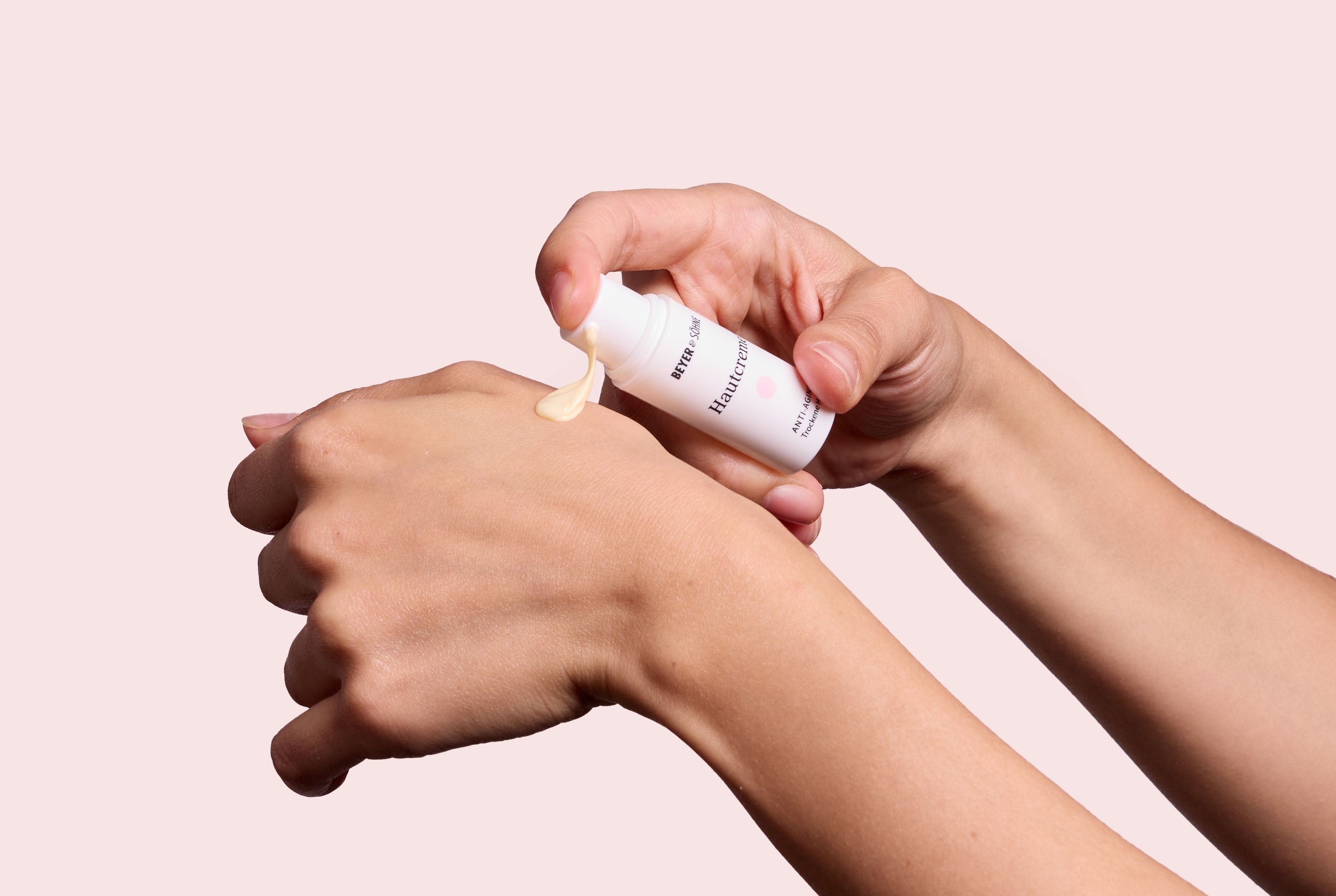 Nahaufnahme von Händen vor rosa Hintergrund, die eine kleine Menge der Beyer & Söhne Hautcreme aus einem niedlichen Tester-Pumpspender auf den Handrücken geben, um die cremige Textur und Anwendung zu demonstrieren.