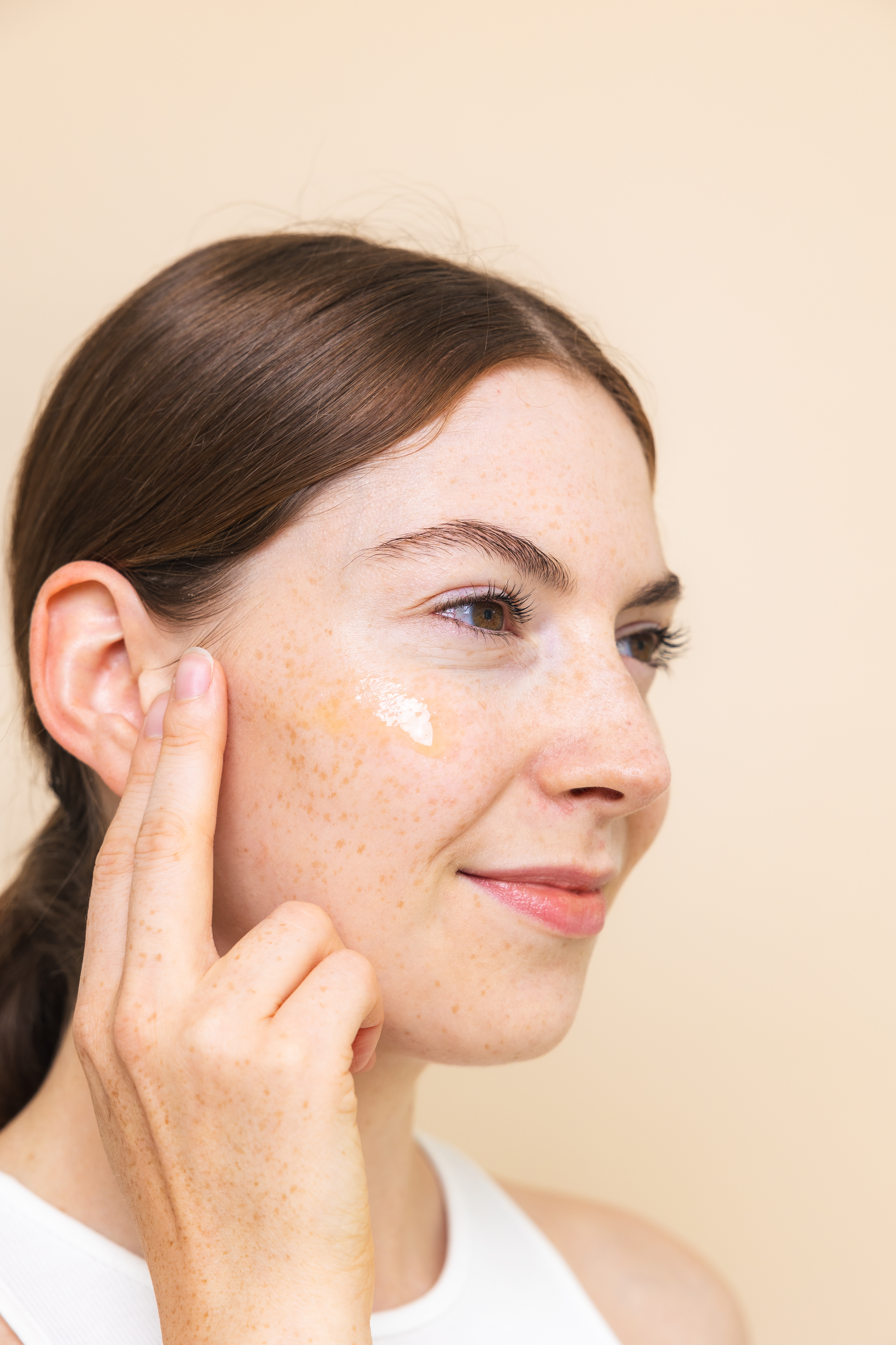 Portraitaufnahme einer jungen, natürlichen Frau mit braunen Pferdeschwanz, die mit einem sanften Lächeln ein Serum auf ihre Wange aufträgt.