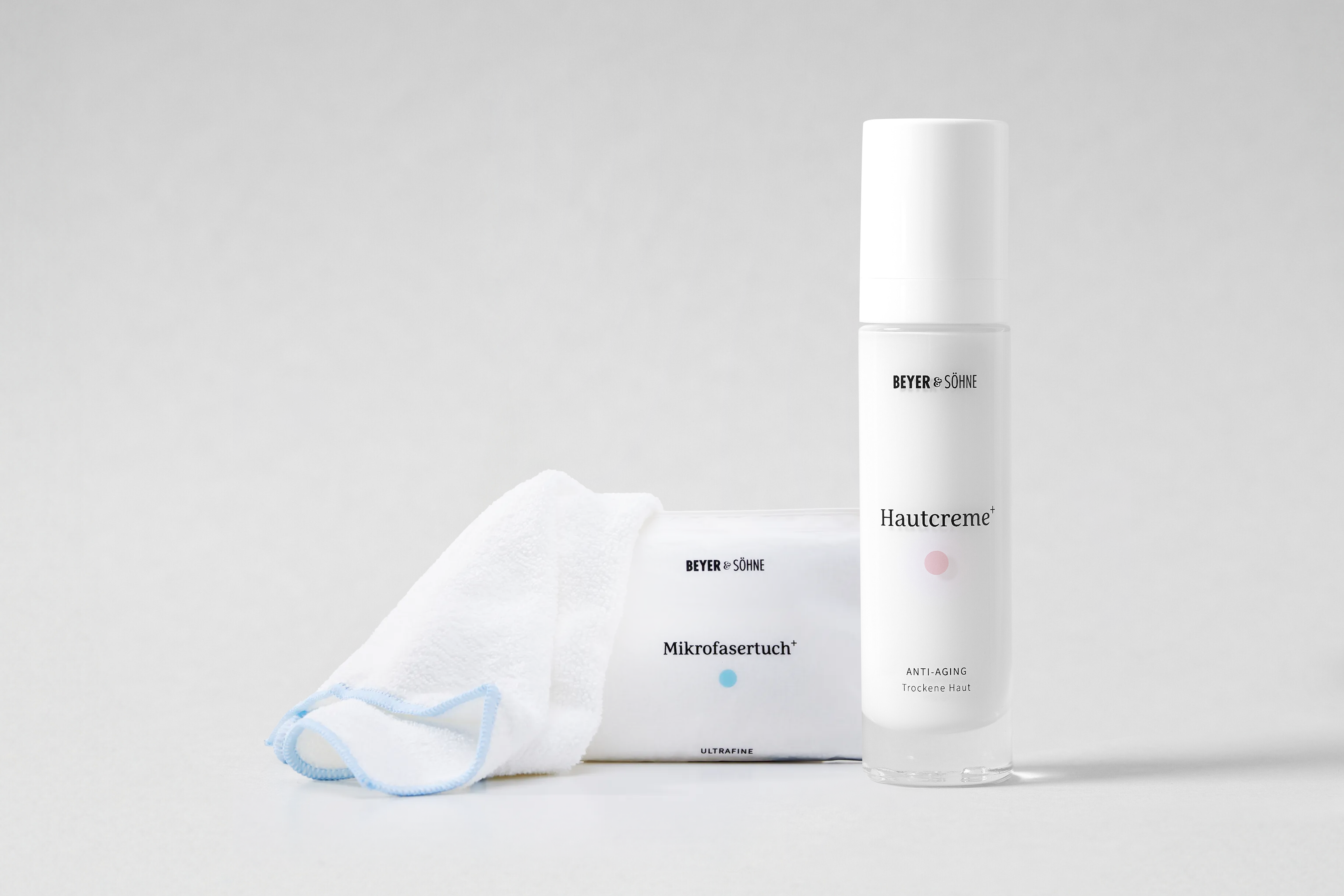 Hautcreme+ Anti-Aging Trockene Haut und Mikrofasertuch+ vor neutral hellem Hintergrund. Die reduzierte Inszenierung betont die hochwertige Pflegeroutine und das minimalistische Produktdesign.