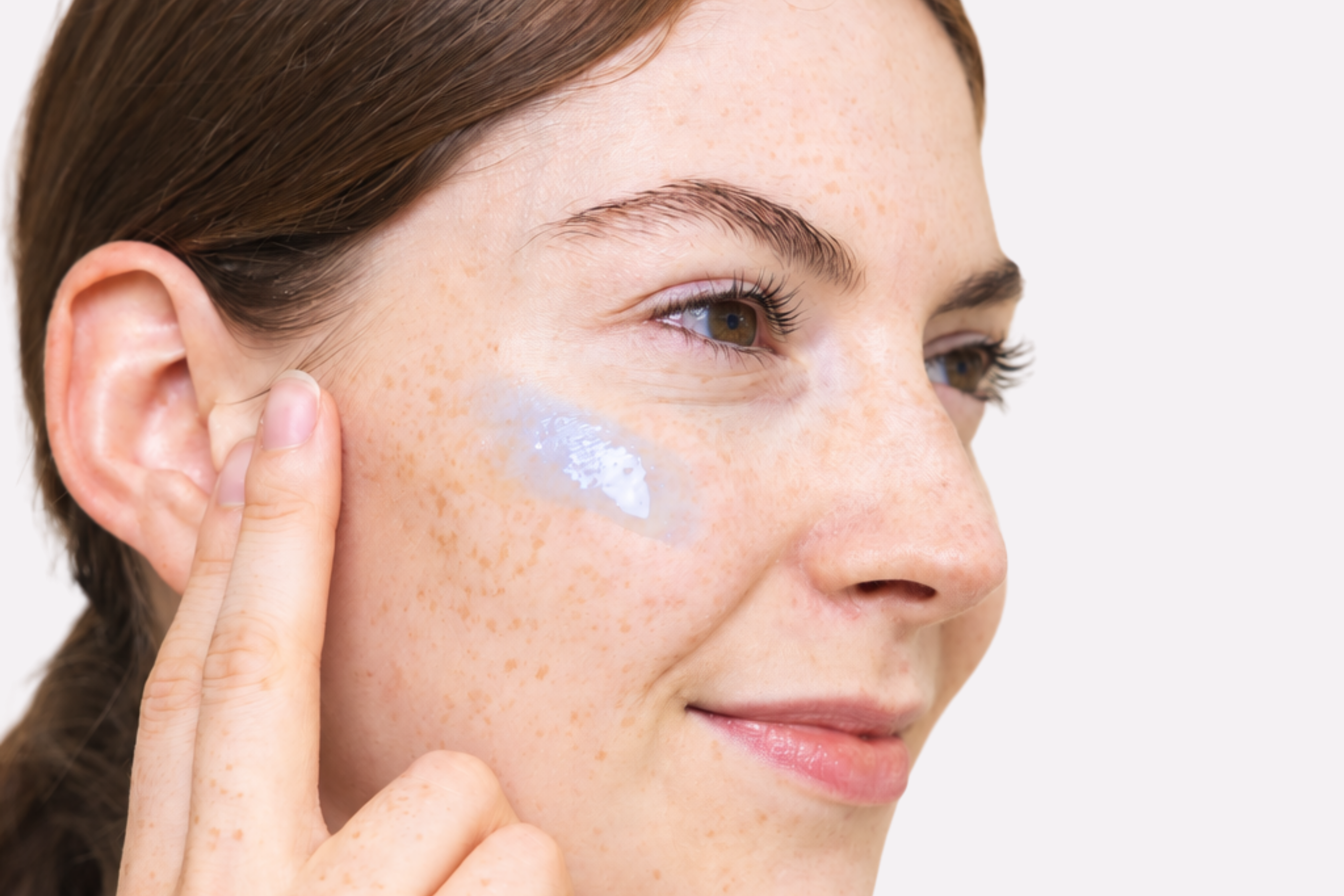 Nahaufnahme einer lächelnden Frau mit natürlichem Hautbild und Sommersprossen trägt die blaue Textur des Peptide Boosters von Beyer & Söhne auf die Wange auf.