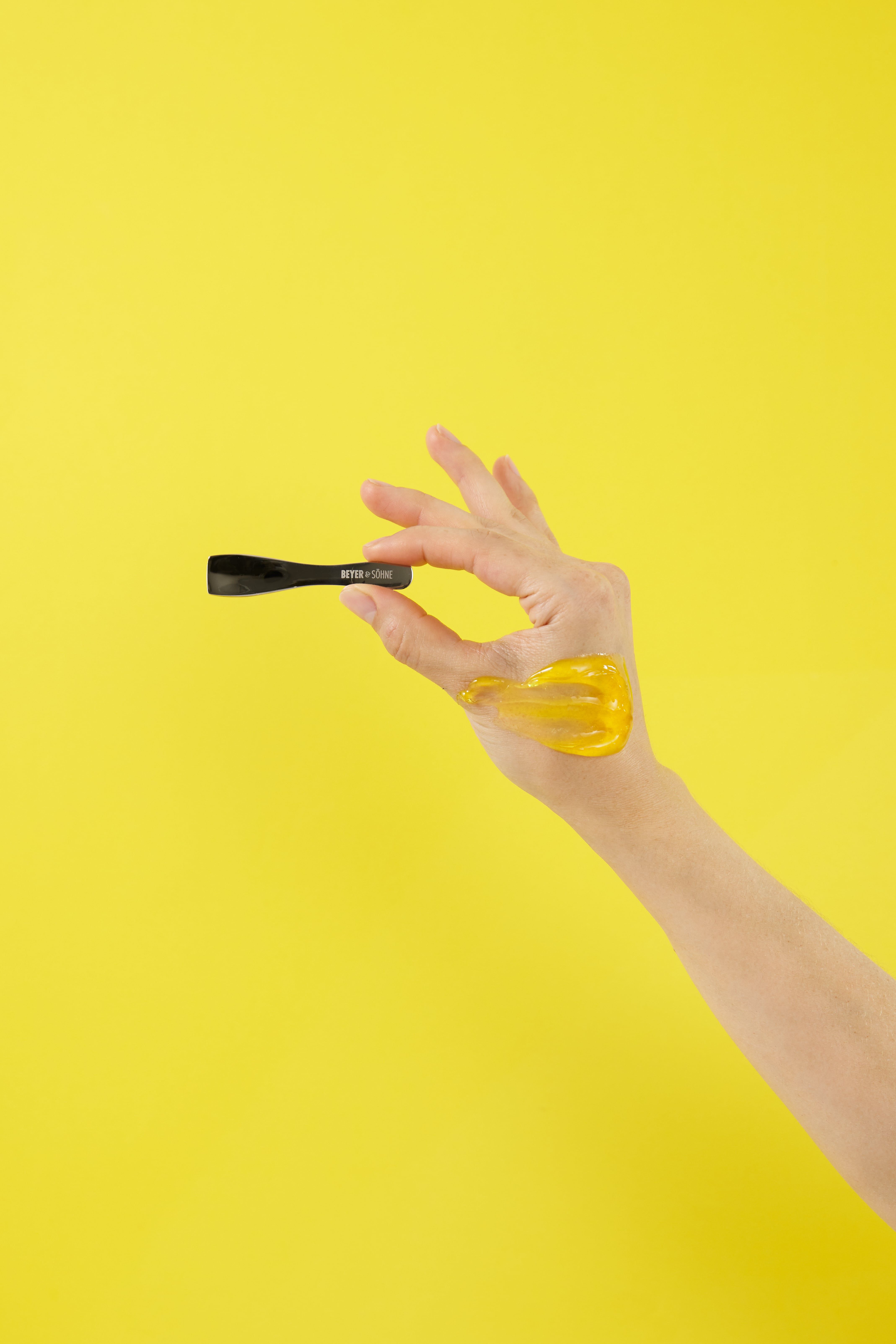 Eine Hand hält einen Spatel aus Edelstahl vor gelbem Hintergrund. Auf der Hand ist der Cleanser verteilt.