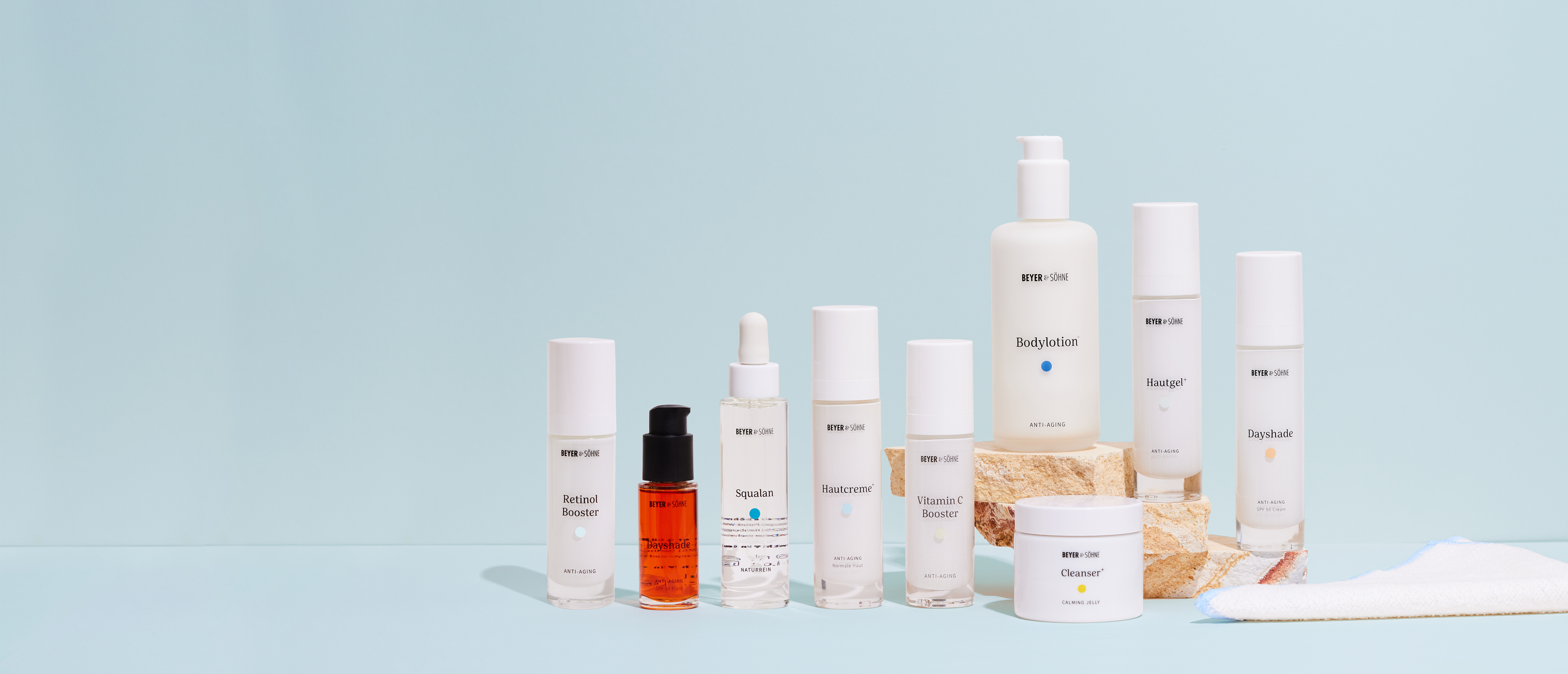 Beyer & Söhne Produktpalette ästhetisch vor einem hellblauen Hintergrund platziert, wobei einige Produkte auf einem beigen Naturstein im Hintergrund stehen