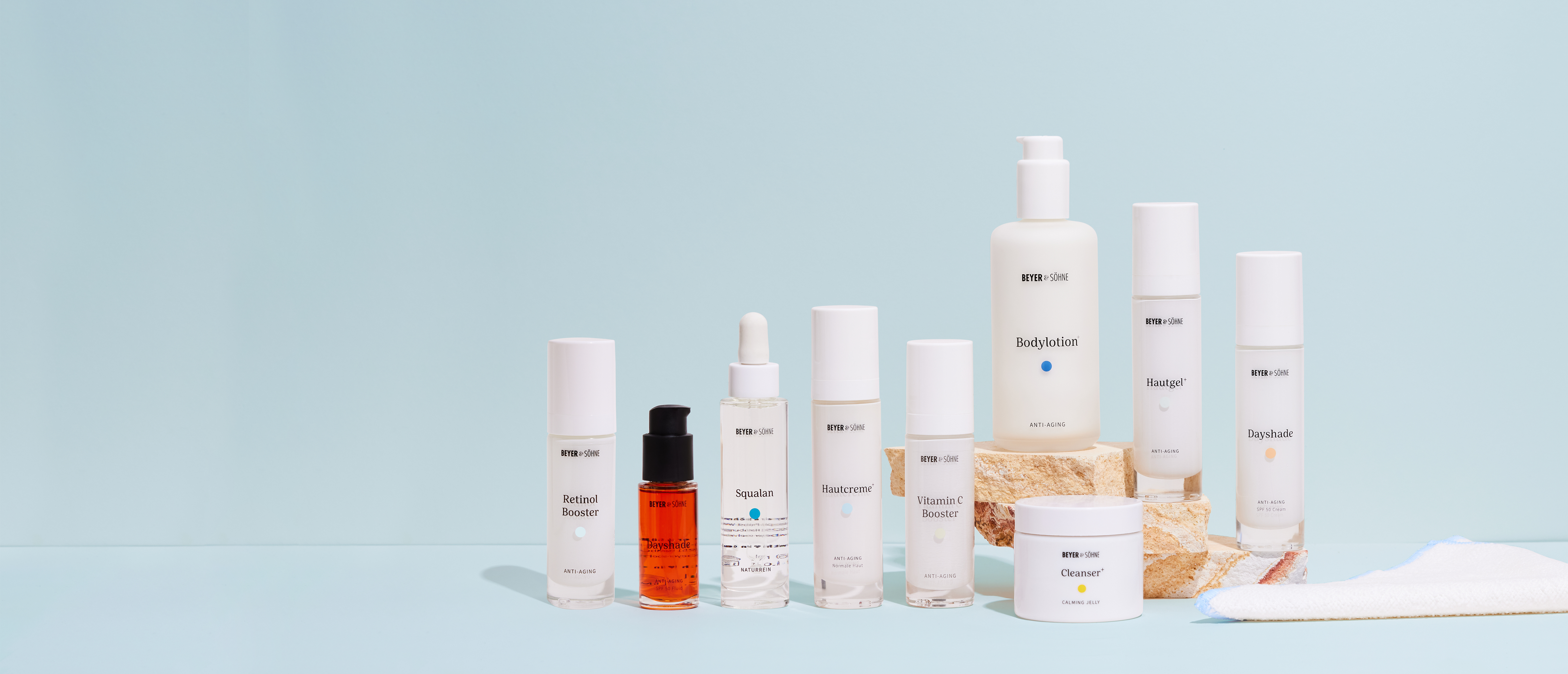 Beyer & Söhne Produktpalette ästhetisch vor einem hellblauen Hintergrund platziert, wobei einige Produkte auf einem beigen Naturstein im Hintergrund stehen