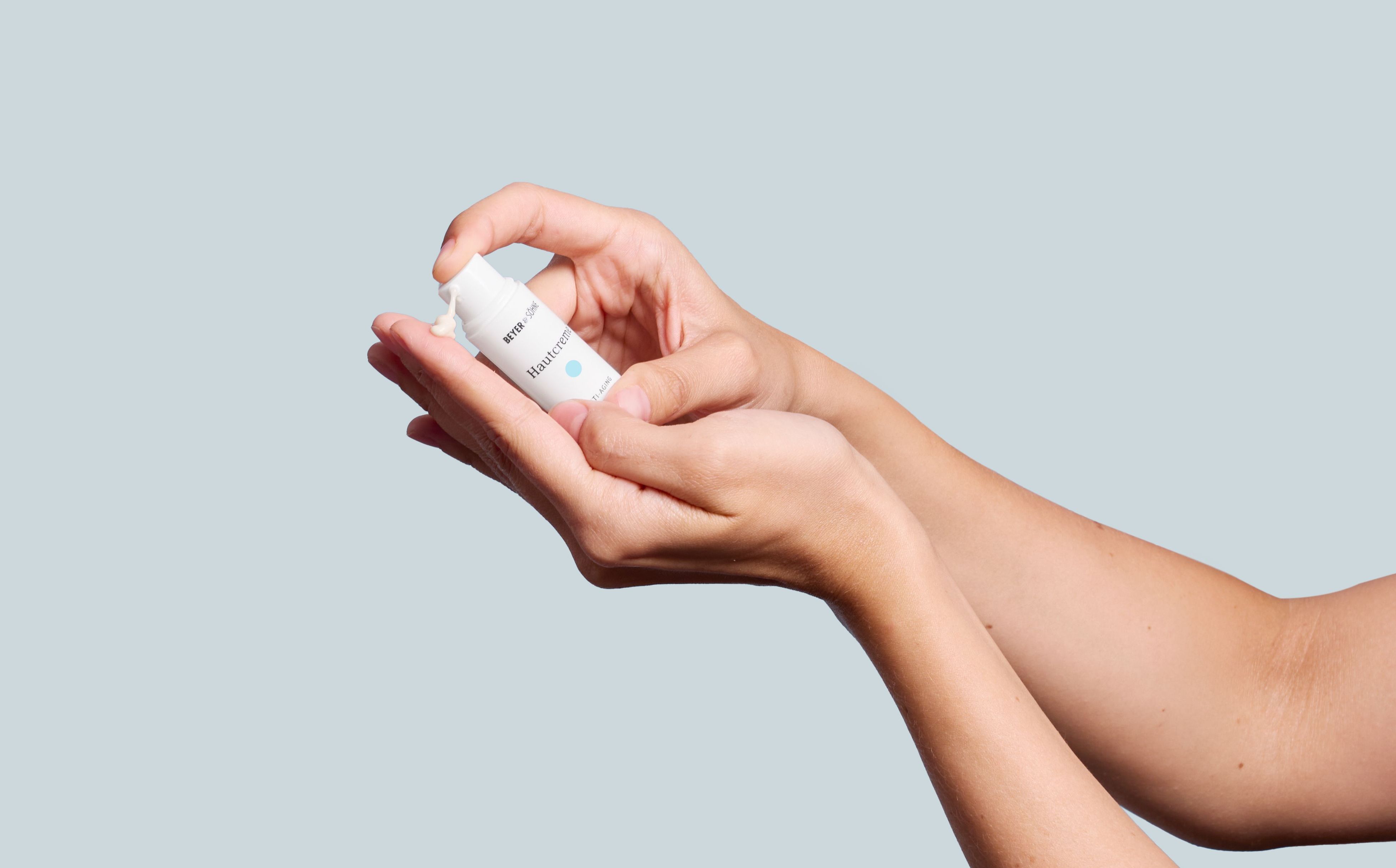 Nahaufnahme von Händen vor hellblauem Hintergrund, die eine kleine Menge der Beyer & Söhne Hautcreme Normal aus einem niedlichen Tester-Pumpspender auf den Handrücken geben, um die cremige Textur und Anwendung zu demonstrieren.