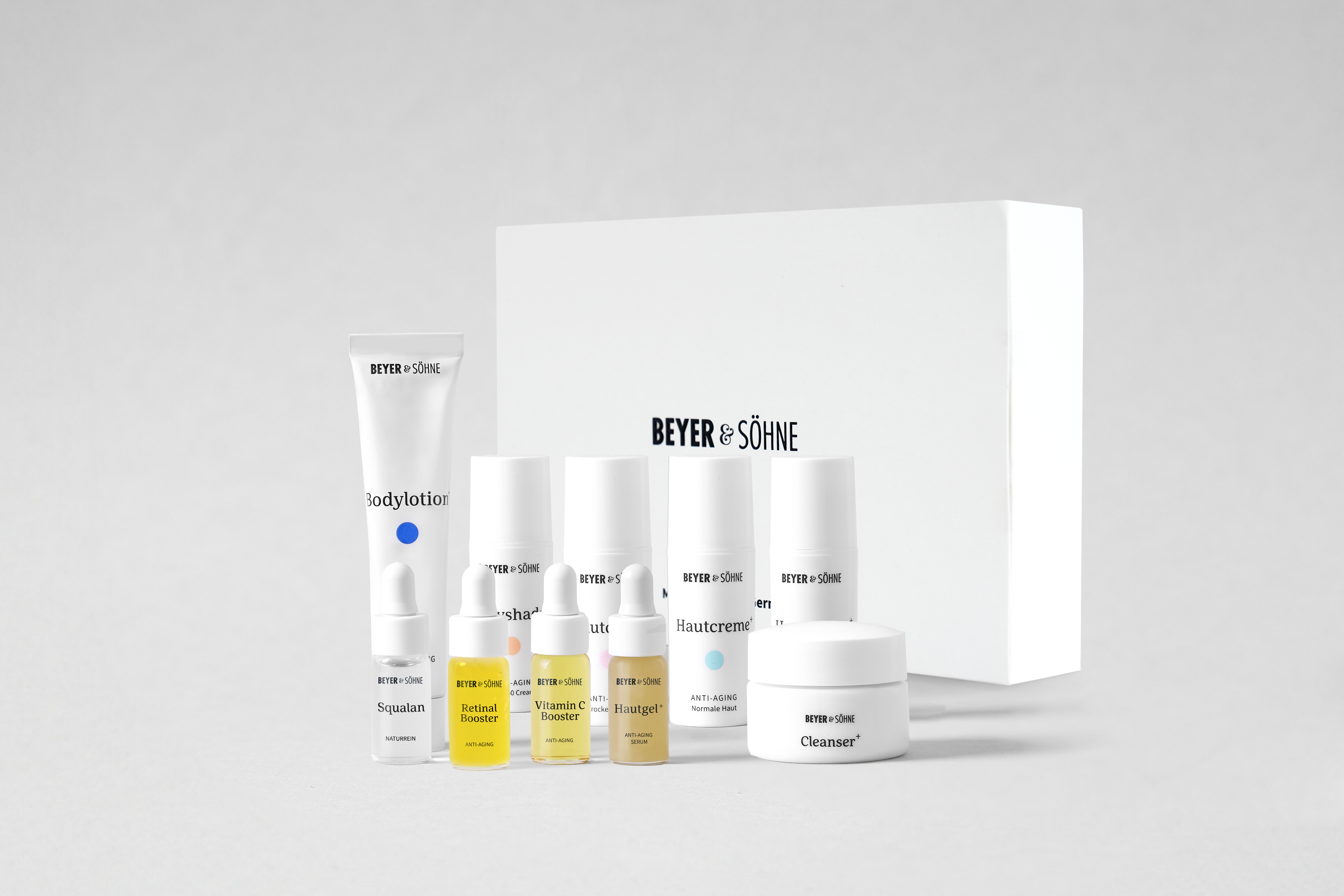 Tester-Set von Beyer & Söhne mit verschiedenen Hautpflegeprodukten als niedliche Minigrößen, darunter Bodylotion, Cleanser, vier Booster (Retinol, Retinal, Vitamin C, Squalan), zwei Hautcremes, ein Sachet mit Dayshade Lippenpflege sowie eine weiße Produktbox im Hintergrund mit Beyer & Söhne Logo