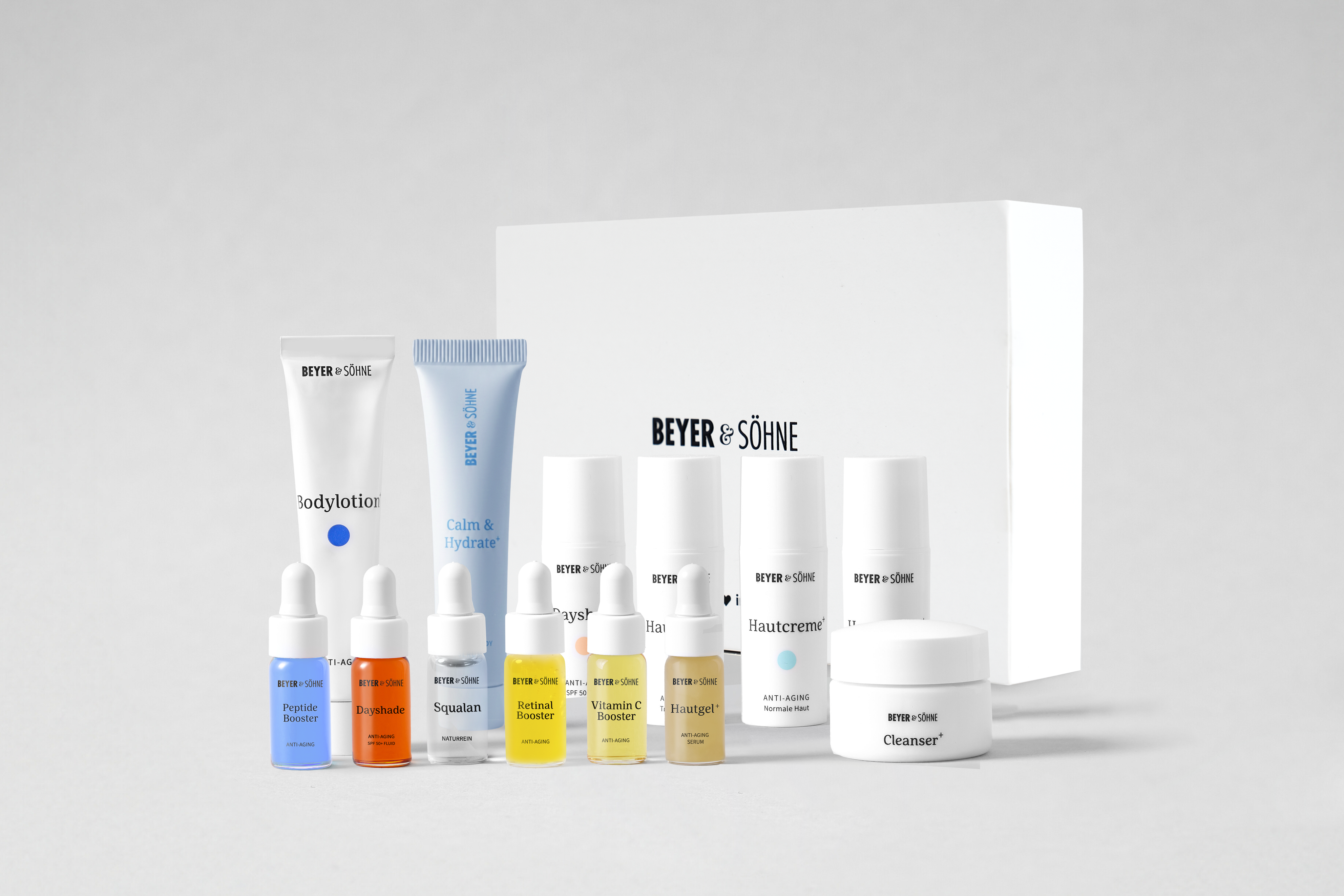 Tester-Set von Beyer & Söhne mit verschiedenen Hautpflegeprodukten in Probiergrößen, darunter Bodylotion+, Calm & Hydrate+, Cleanser+, Dayshade Fluid LSF 50, Dayshade Cream LSF 50, Squalan, Vitamin C Booster, Retinal Booster, Peptide Booster, Hautgel+ sowie Hautcreme+ Leicht, Normale Haut und Trockene Haut. Im Hintergrund die weiße Tester-Set-Box mit Beyer & Söhne Logo.