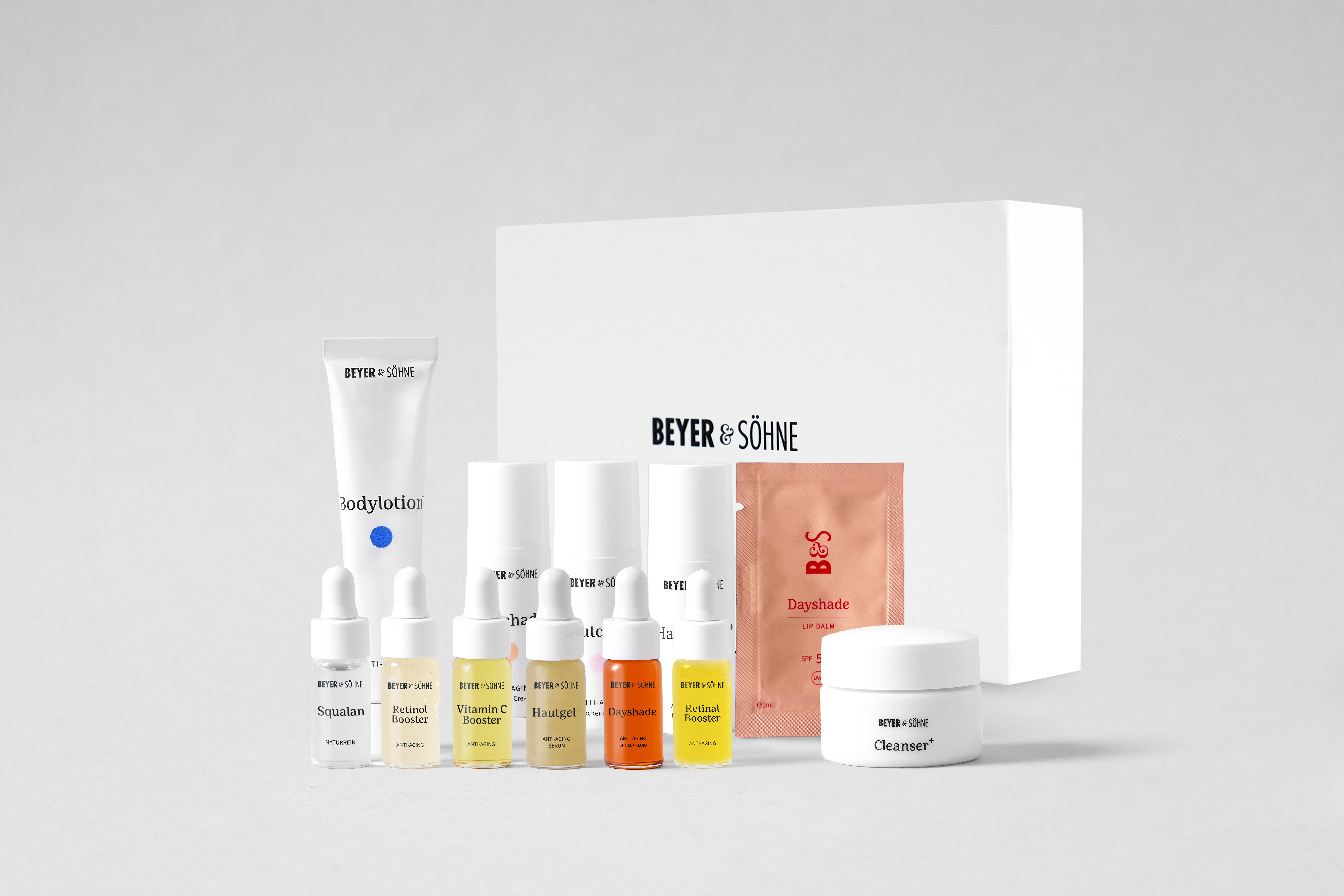 Tester-Set von Beyer & Söhne mit verschiedenen Hautpflegeprodukten als niedliche Minigrößen, darunter Bodylotion, Cleanser, vier Booster (Retinol, Retinal, Vitamin C, Squalan), zwei Hautcremes, ein Sachet mit Dayshade Lippenpflege sowie eine weiße Produktbox im Hintergrund mit Beyer & Söhne Logo