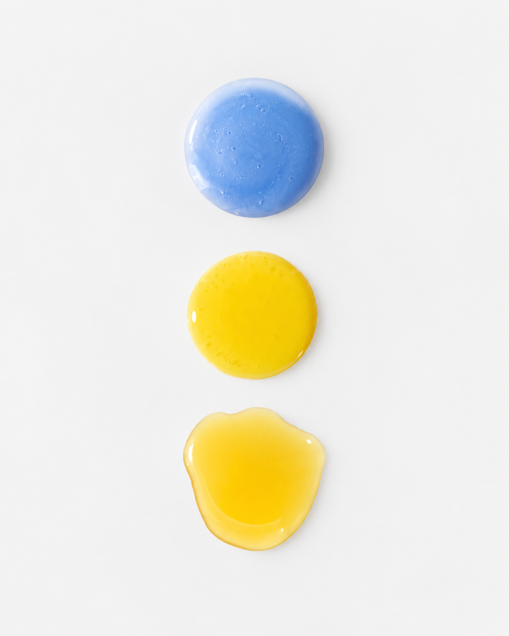 Drei Serum-Texturen auf grauem Hintergrund untereinander. Oben die blaue Peptide Booster Textur, mittig die gelbe Textur des Retinal Boosters und unten die ebenfalls gelbe aber flüssigere Textur des Vitamin C Boosters