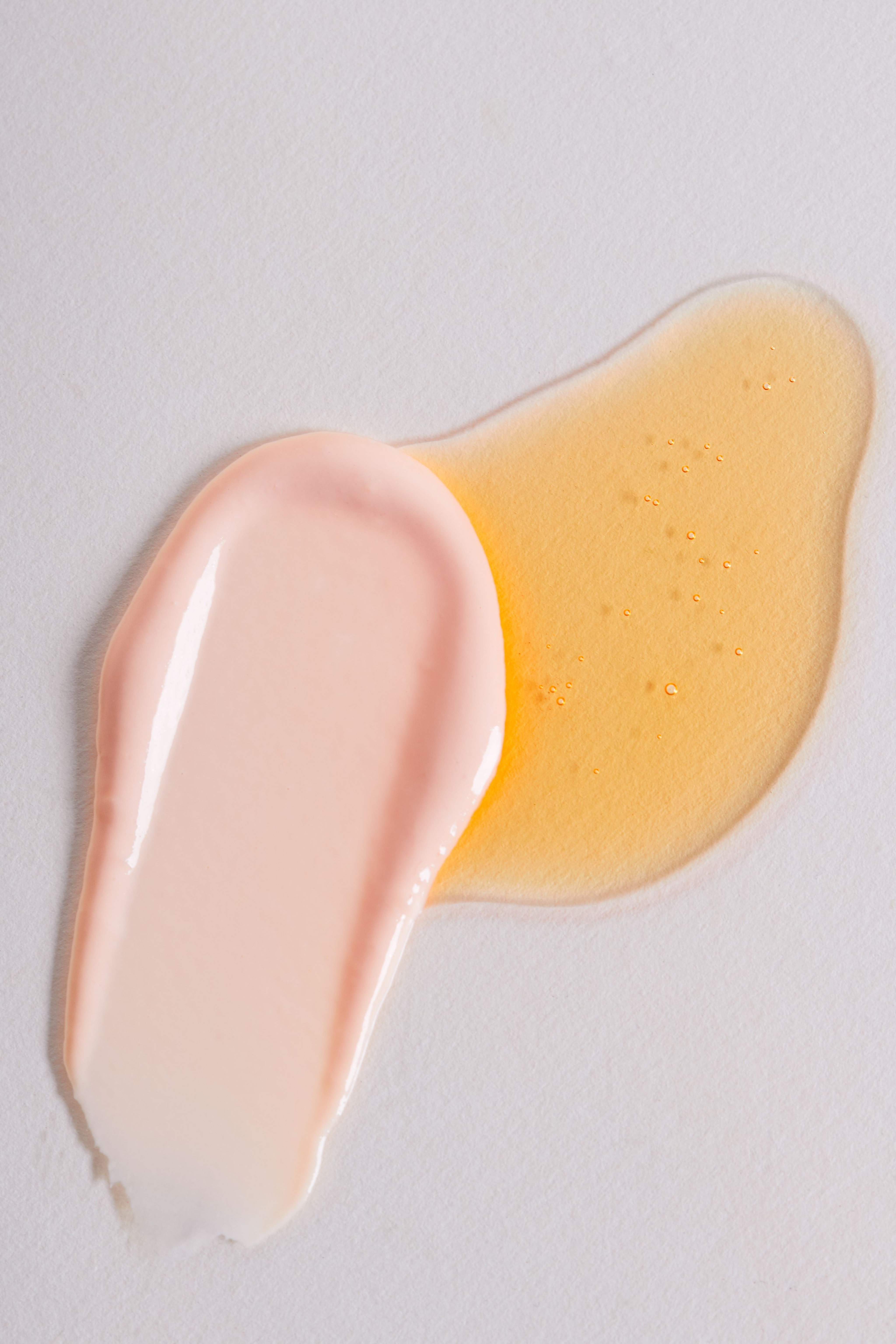 Nahaufnahme von zwei Produkttexturen: Links die cremige, rosafarbene Dayshade SPF 50 Cream, rechts das rötliche, flüssige Dayshade SPF 50+ Fluid