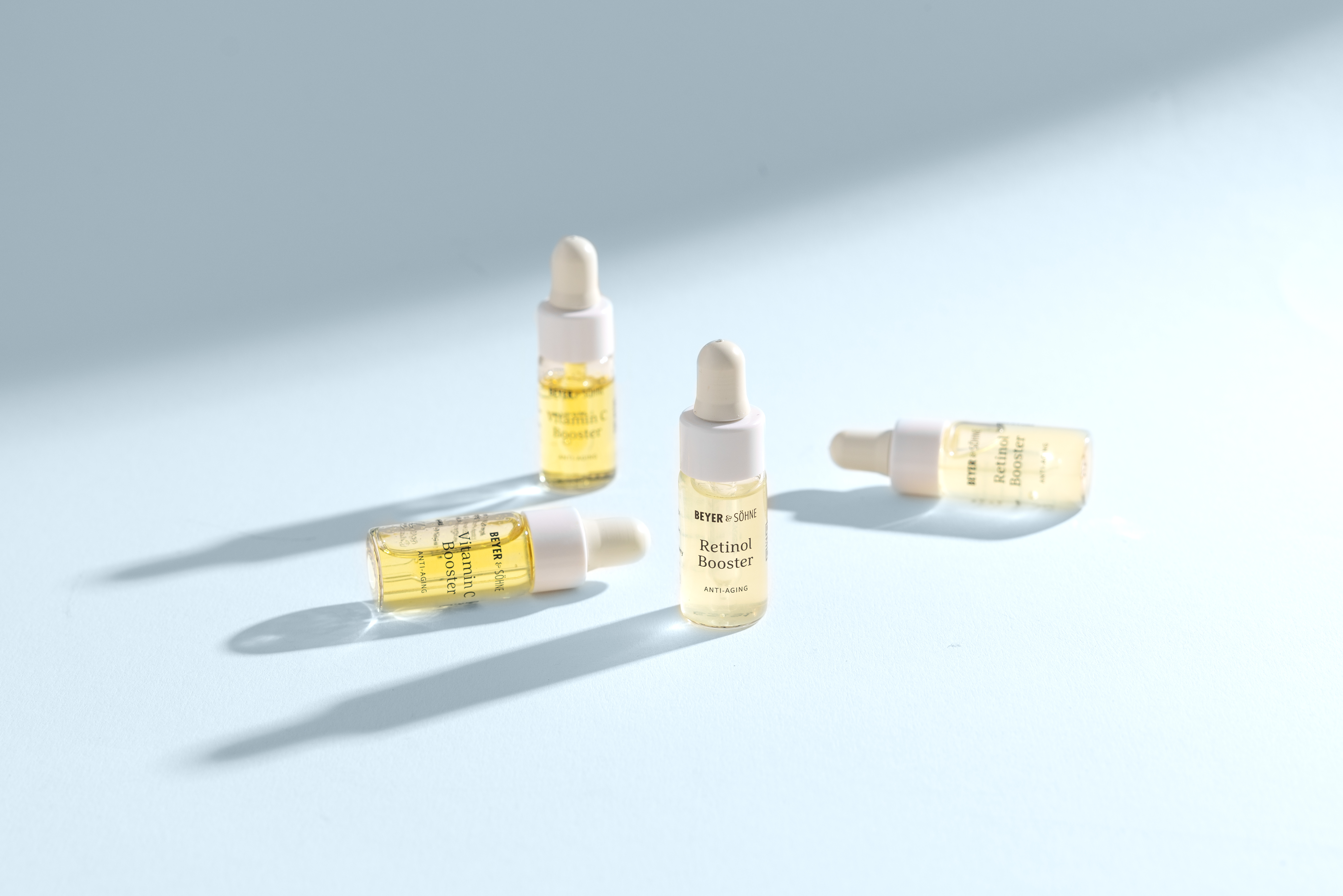 Vier kleine Flaschen mit Pipette mit der Aufschrift Retinol Booster und Vitamin C von Beyer & Söhne vor hellblauem Hintergrund. Licht strahlt sanft von der Seite durch die hellgelbe und zitronengelbe Textur.