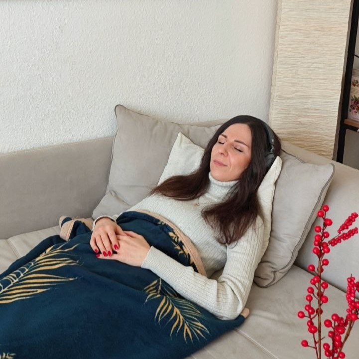 Frau mit Kopfhörern liegt entspannt auf dem Sofa, lächelt sanft und hört eine Imaginationsreise.