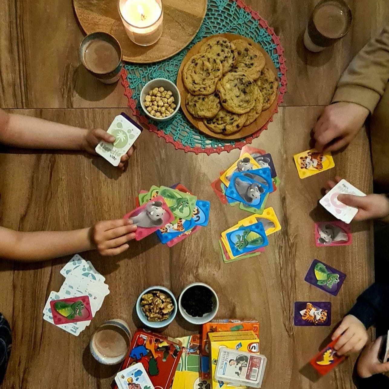 Eine Familie spielt ein Kartenspiel in gemütlicher Atmosphäre an einem Holztisch, umgeben von Keksen, Nüssen, Kakao und einer brennenden Kerze.