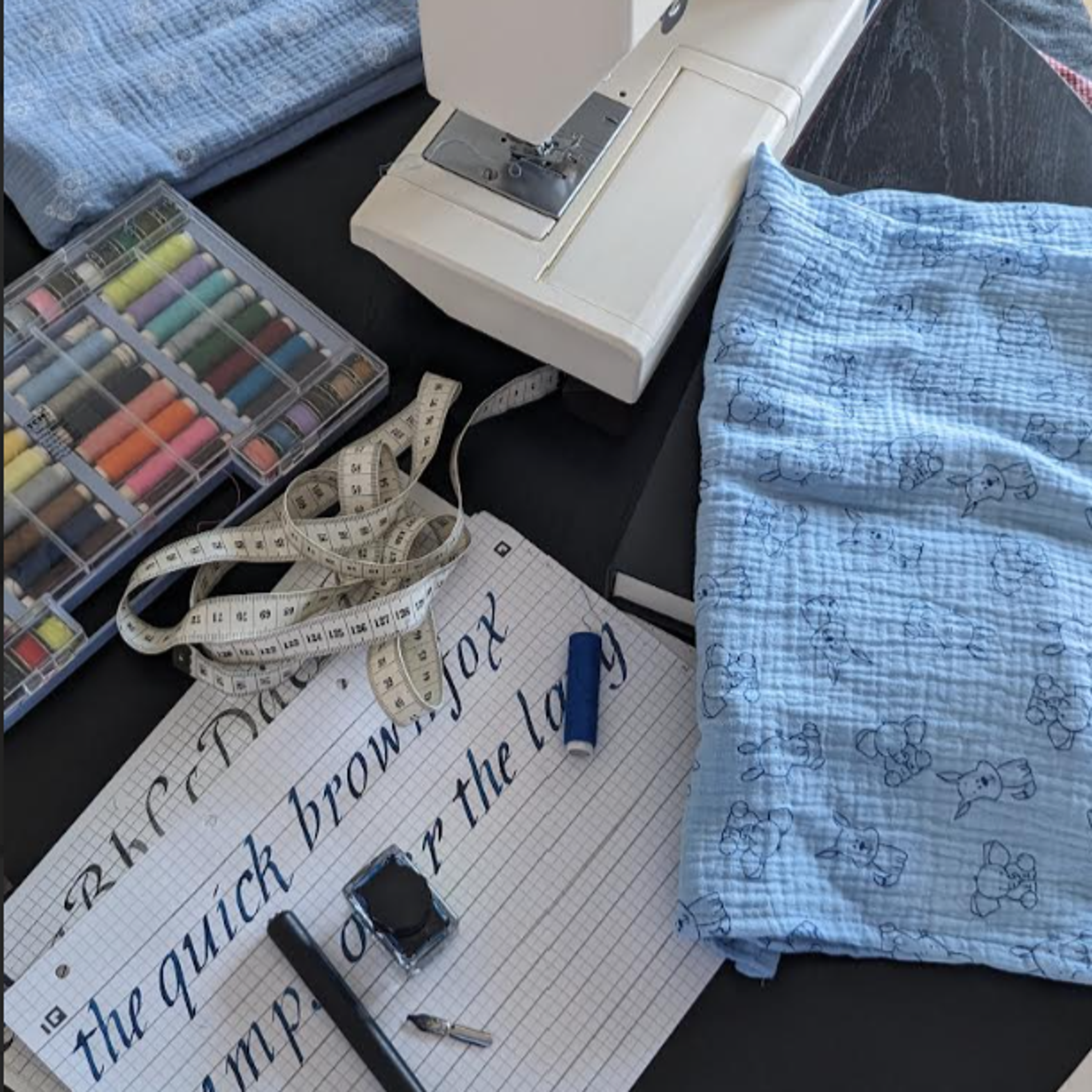 Ein kreativer Arbeitsplatz mit einer Nähmaschine, blauem Stoff mit niedlichen Tiermustern, Nähzubehör, einem Maßband und einem Kalligrafie-Übungsblatt 