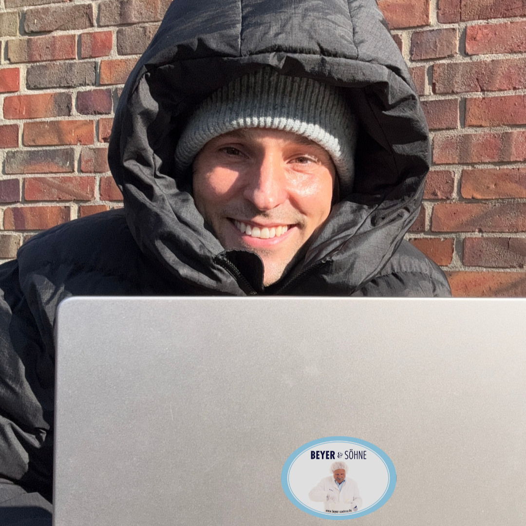 Robby Beyer sitzt lächelnd in dicker Winterjacke und Mütze mit seinem Laptop draußen vor einer Backsteinwand und die Sonne strahlt ihm ins Gesicht.