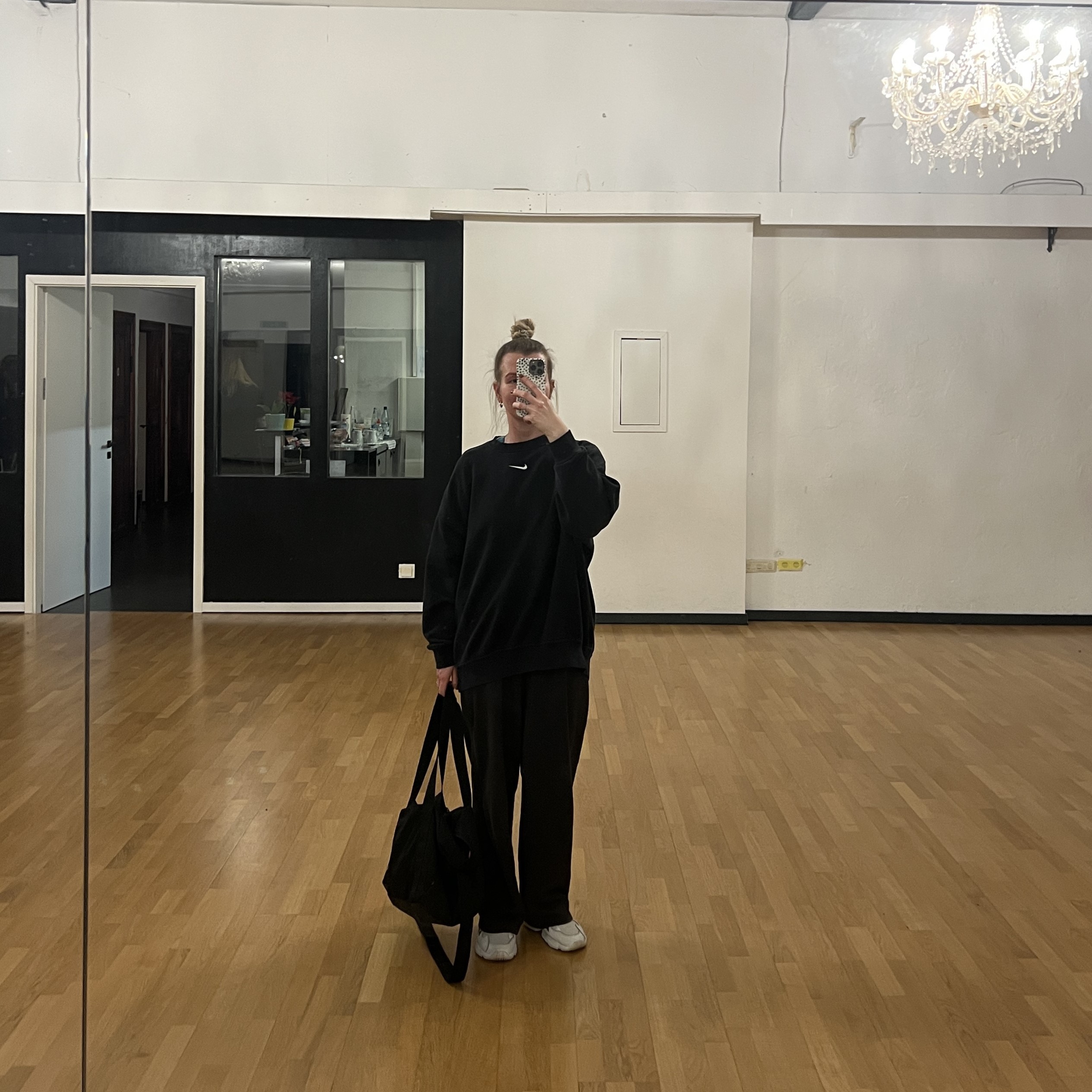 Sarah macht in einem schwarzen Sport-Outfit ein Spiegel-Selfie in einem Tanzstudio mit Holzboden und Kronleuchter. Sie hält eine schwarze Sporttasche in der Hand.