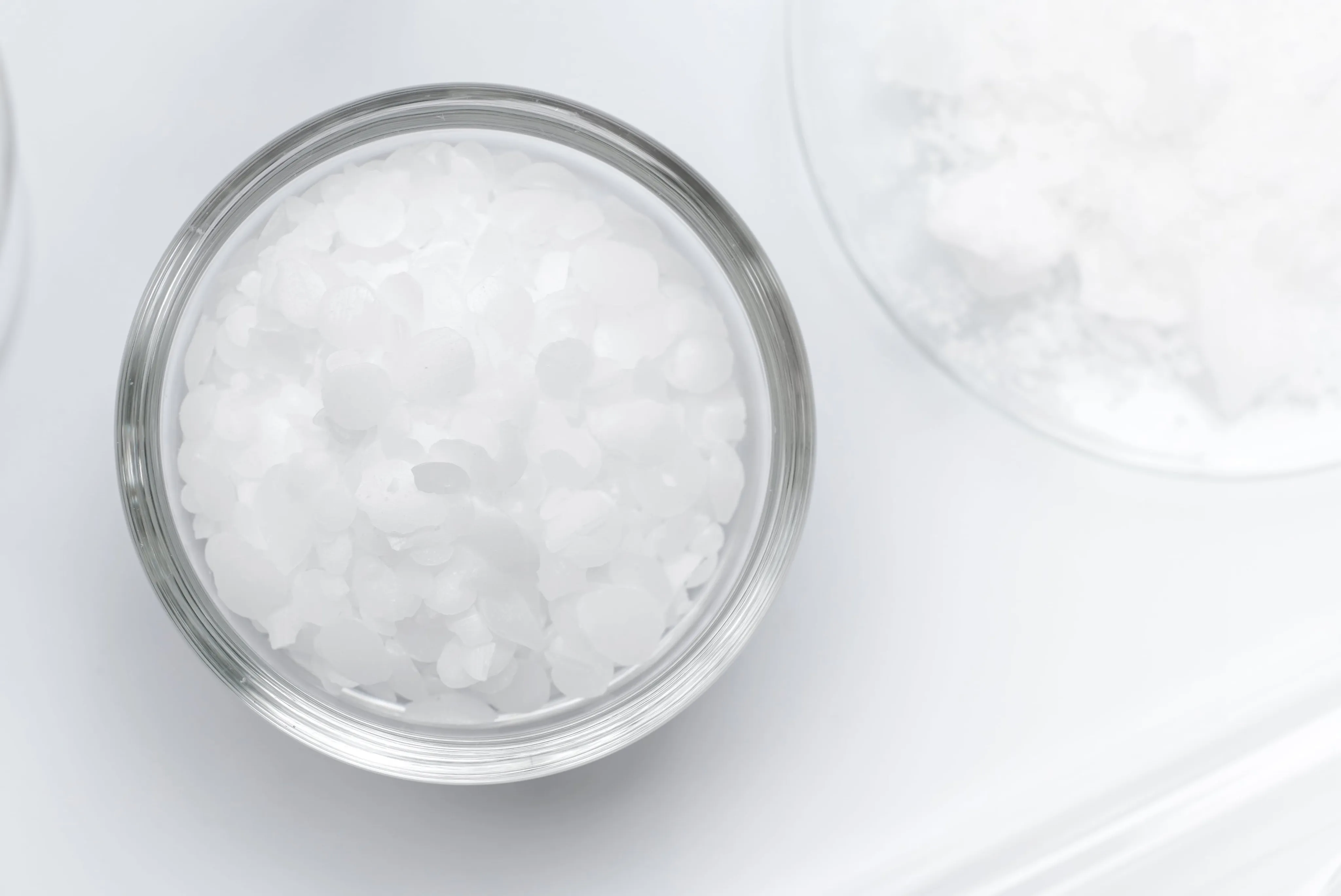 Nahaufnahme eines Glasschälchens mit Sucrose Palmitate in Form von kleinen, weißen, flockigen Kristallen vor einem sauberen, minimalistischen weißen Hintergrund.
