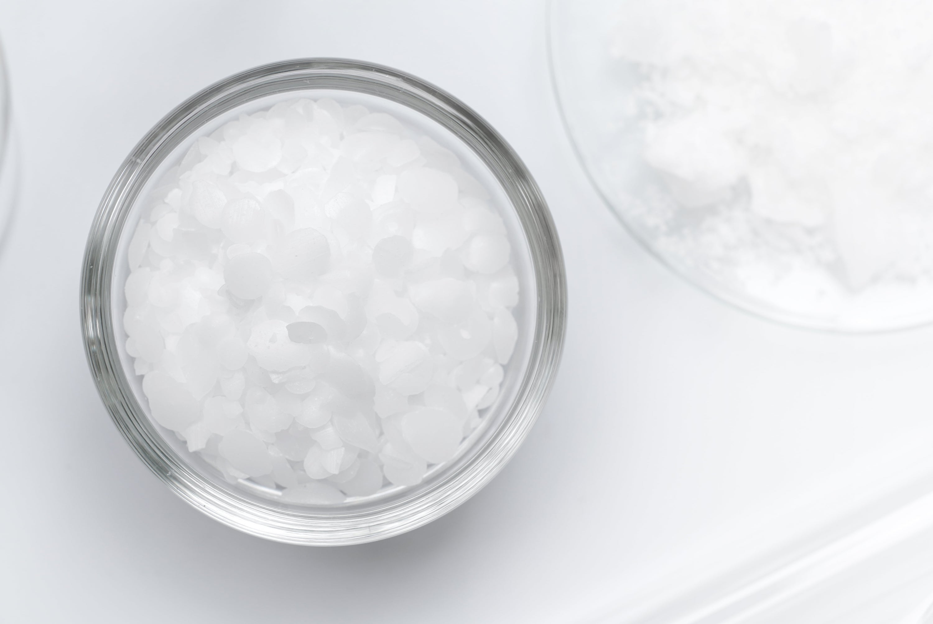 Nahaufnahme eines Glasschälchens mit Sucrose Palmitate in Form von kleinen, weißen, flockigen Kristallen vor einem sauberen, minimalistischen weißen Hintergrund.