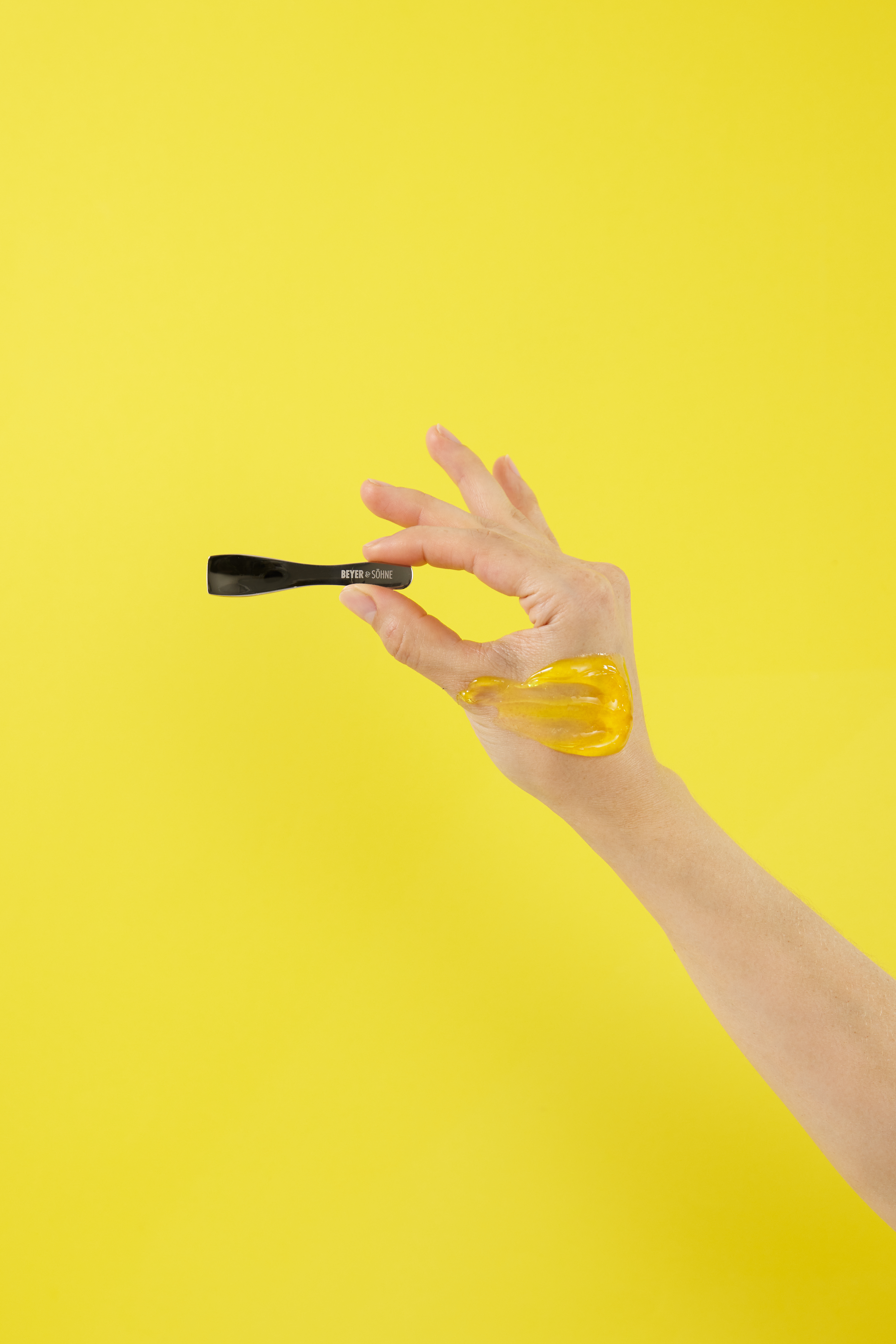 Hand mit einem eleganten Edelstahl-Spatel von Beyer & Söhne und goldfarbenem Gel auf dem Handrücken vor einem leuchtend gelben Hintergrund. 