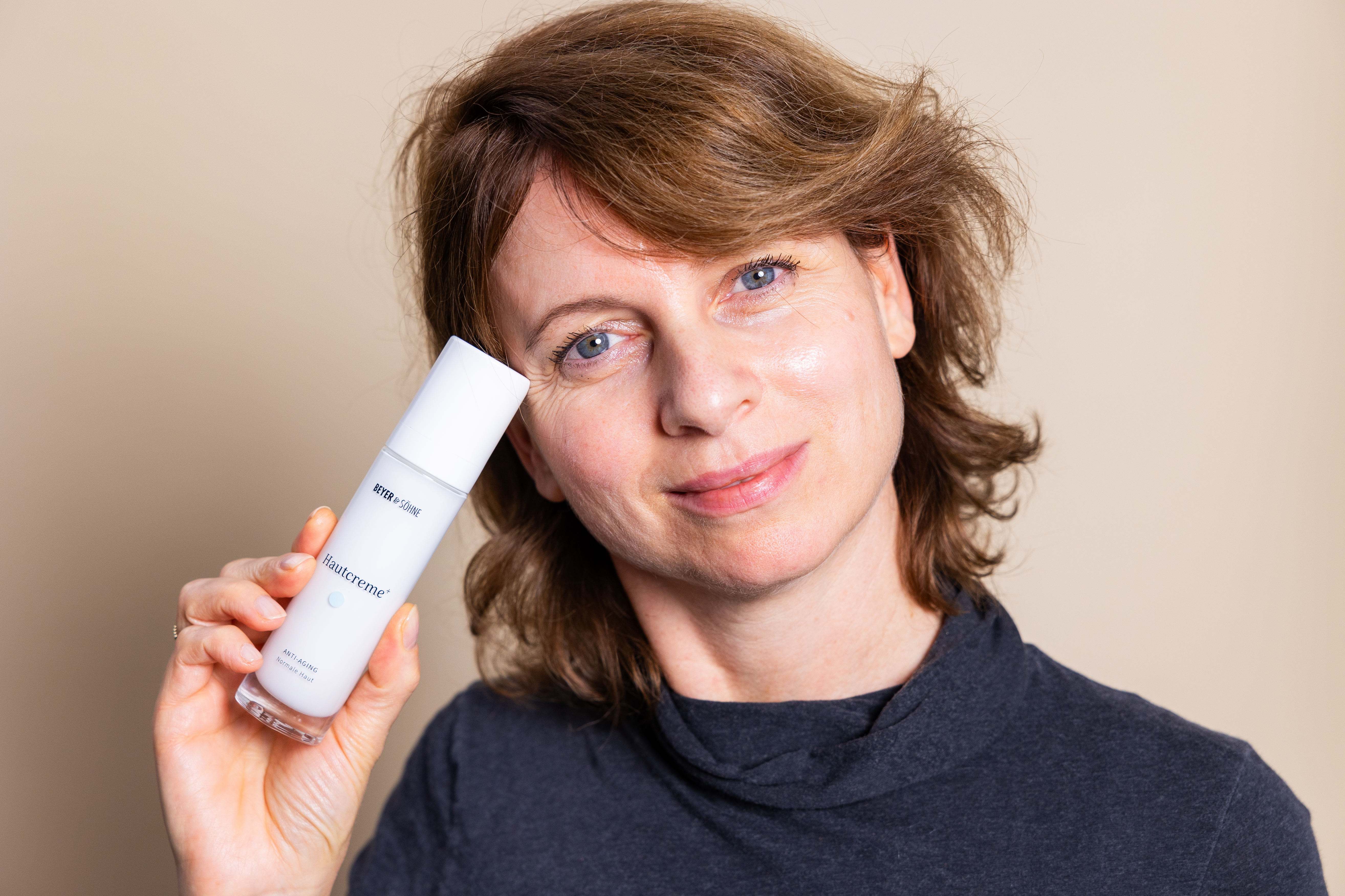 Portrait Aufnahme einer natürlichen Frau Mitte 40 mit sanften Lächeln und einer strahelnden Haut, die zufrieden in die Kamera schauend die Hautcreme+ Normal gegen ihr Gesicht hält.