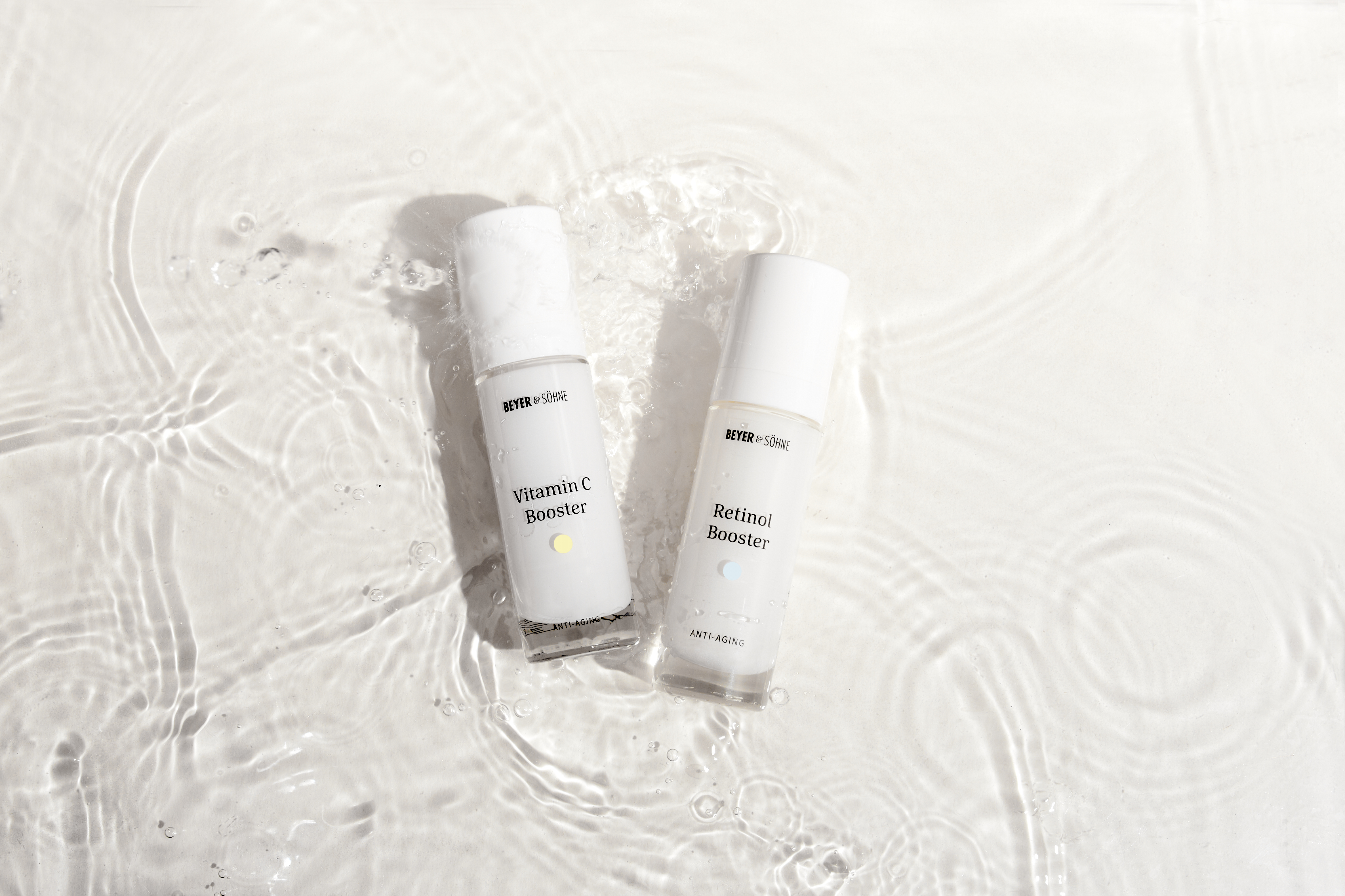 Vitamin C Booster und Retinol Booster von Beyer & Söhne in einer Wasserinszenierung: Beide Anti-Aging-Produkte liegen in klarem, sanft bewegtem Wasser auf weißem Untergrund. 