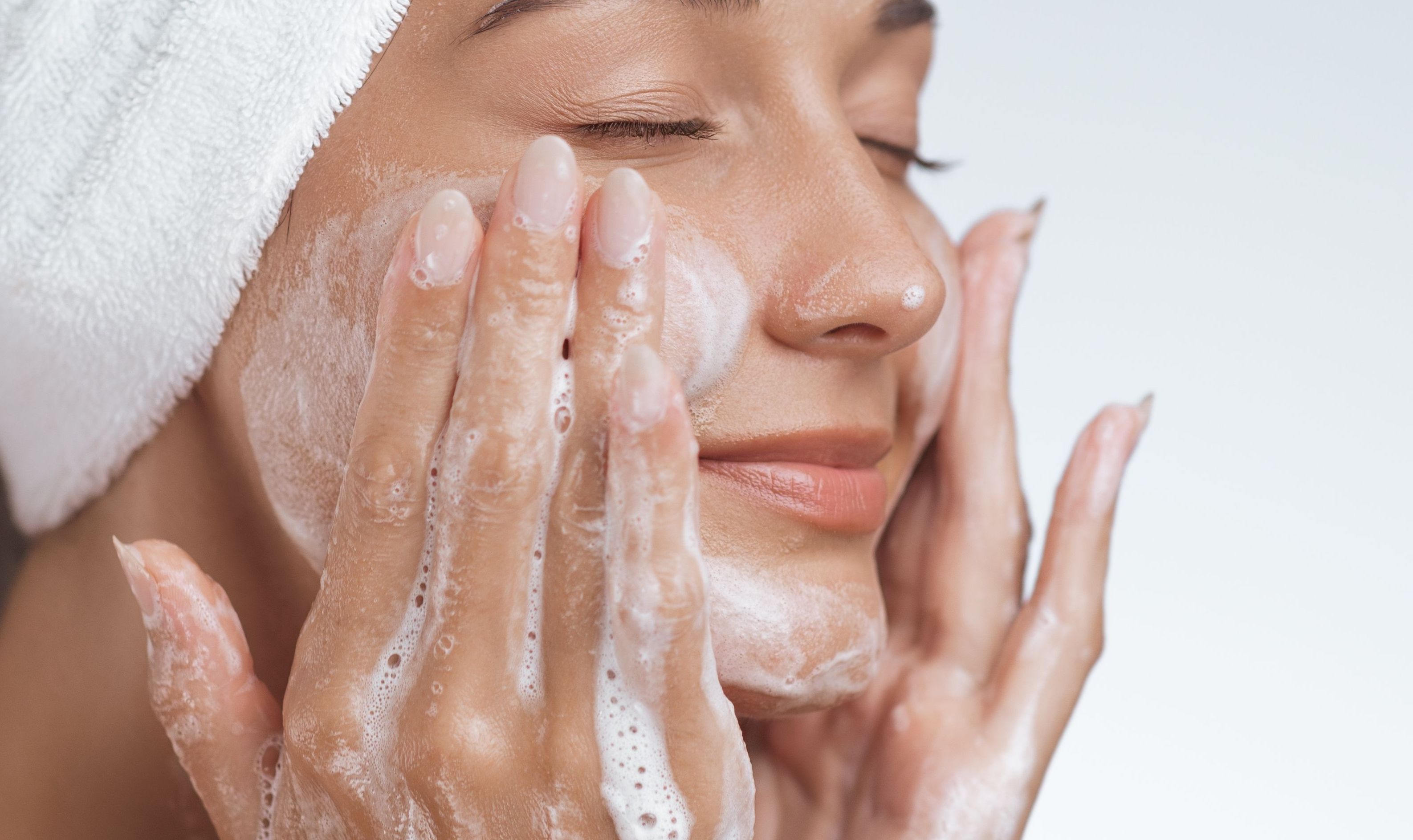 Eine natürlich aussehende Frau im Halbprofil vor einem neutralen, hellgrauen Hintergrund reinigt ihr Gesicht mit einem entspannten Gesichtsausdruck und geschlossenen Augen